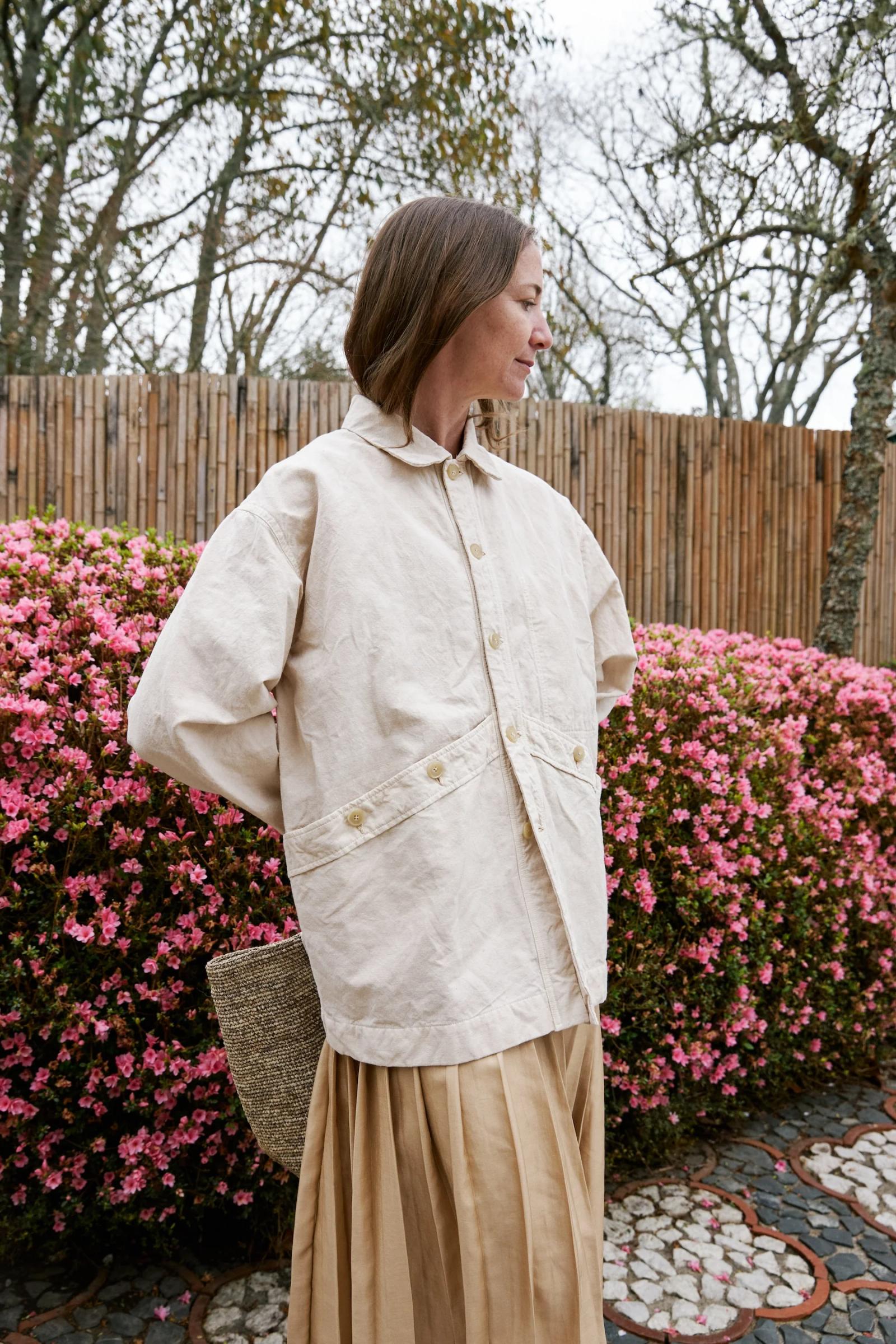 Loose fitting cream jacket is worn over a pale pleated skirt with standing against pink flowers.