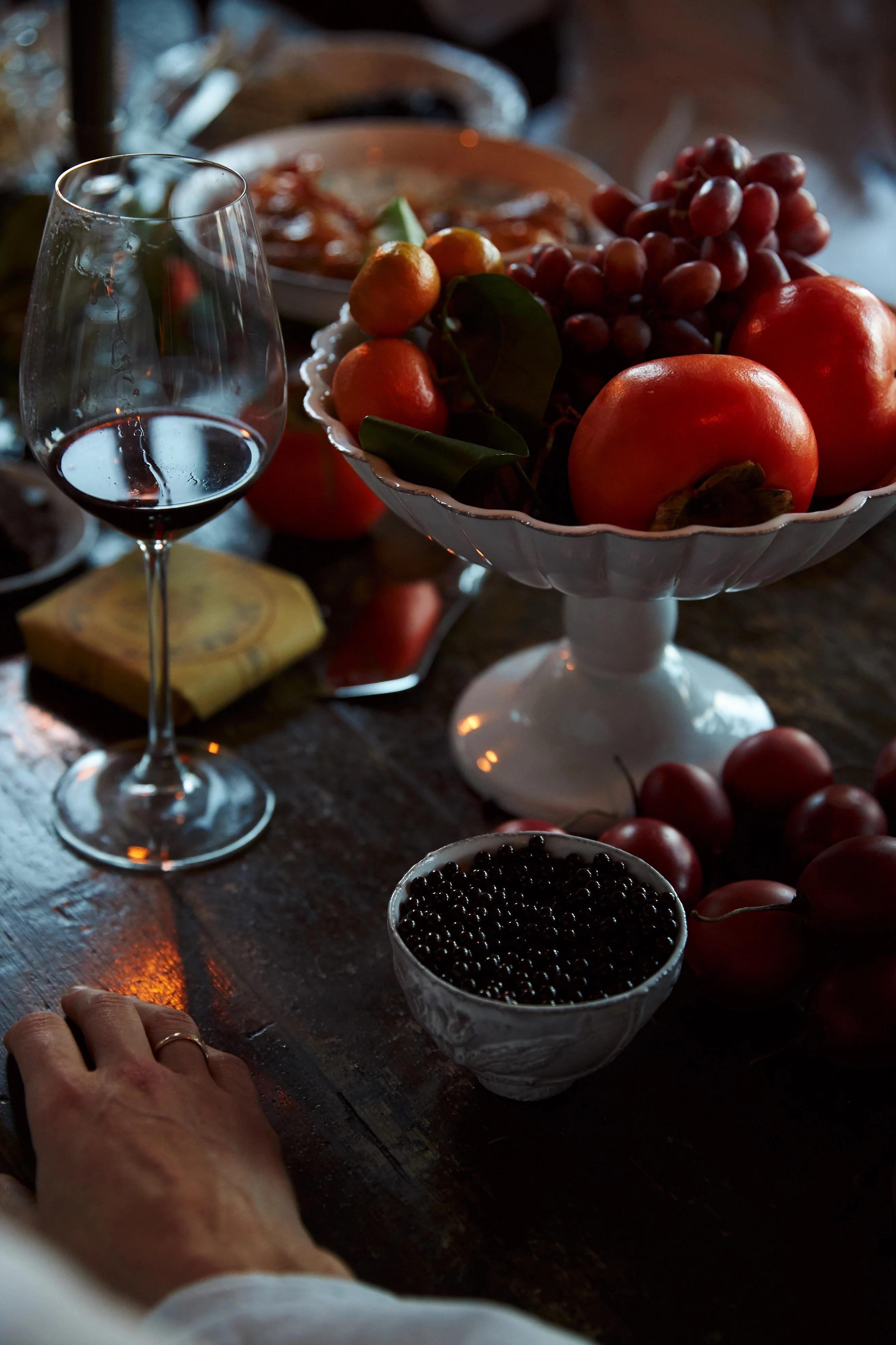 Table with wine, fruit bowls, and vegetables. Person's hand visible at edge of frame.