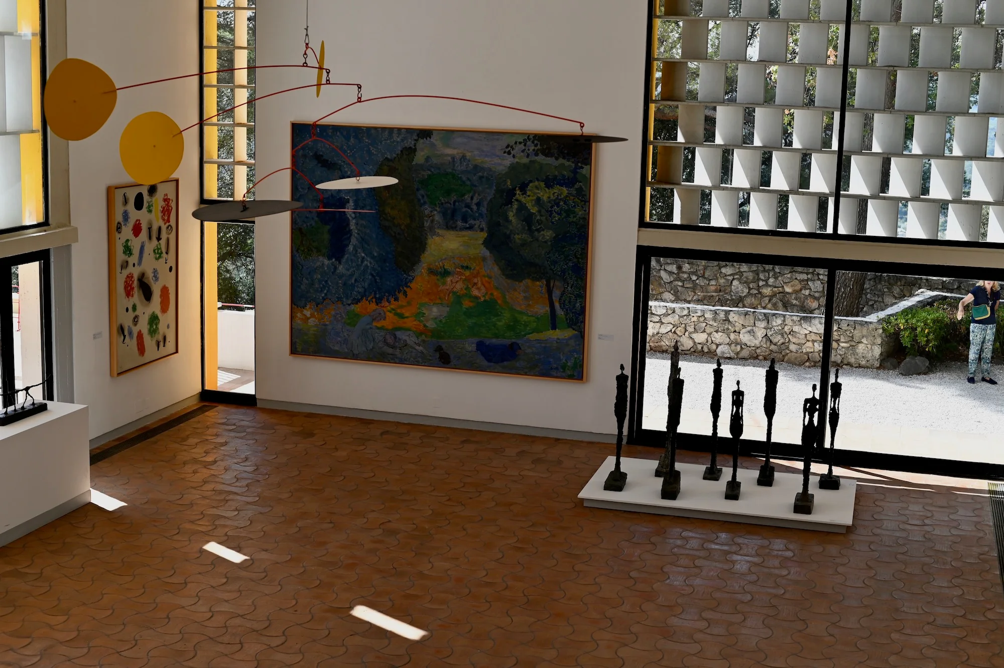 Gallery interior with paintings, hanging mobile, and thin sculptures. Bright light through checkered windows.