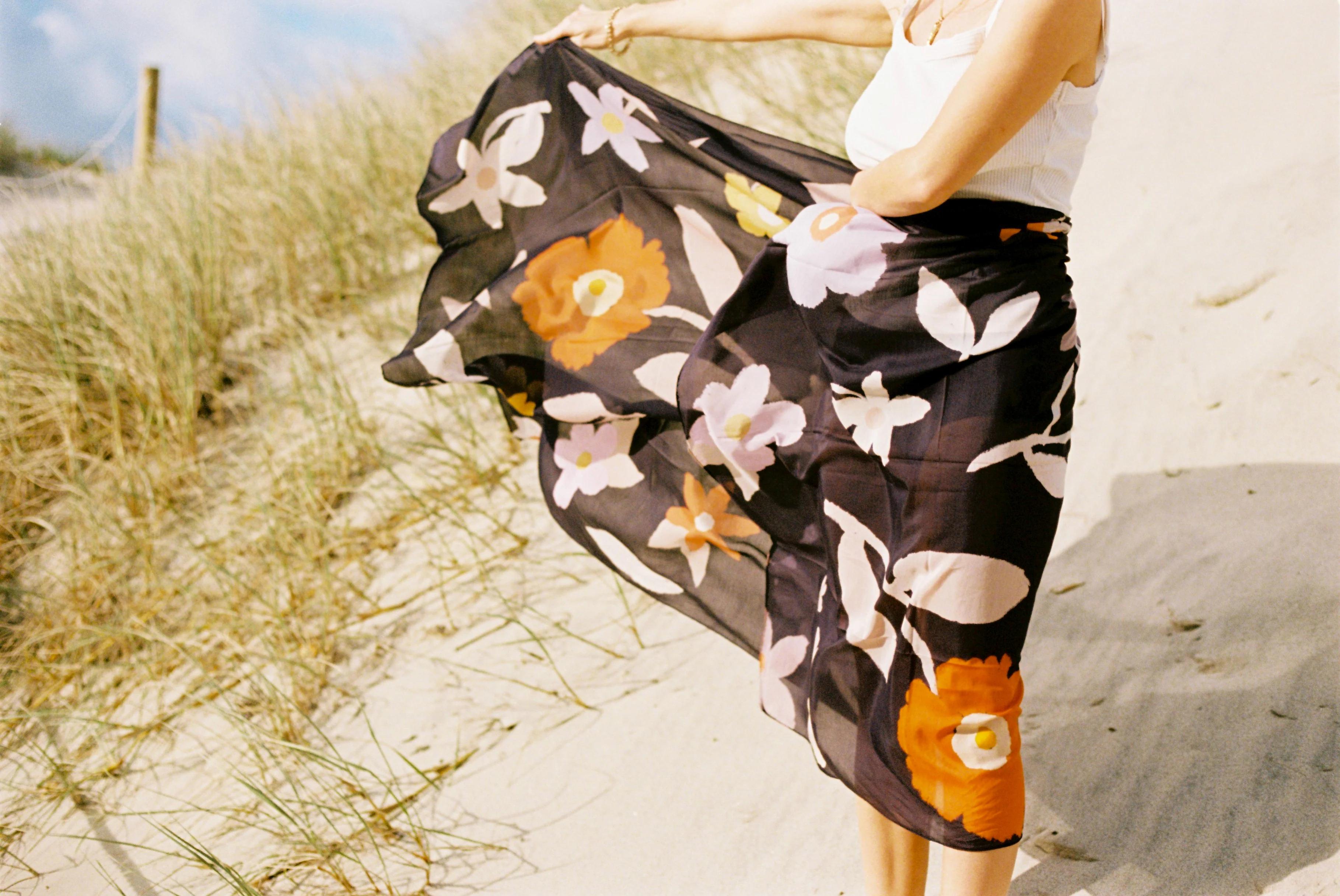 Person on sandy beach holding flowing black fabric with large floral prints. Head out of frame. Native plants and clear sky in background.