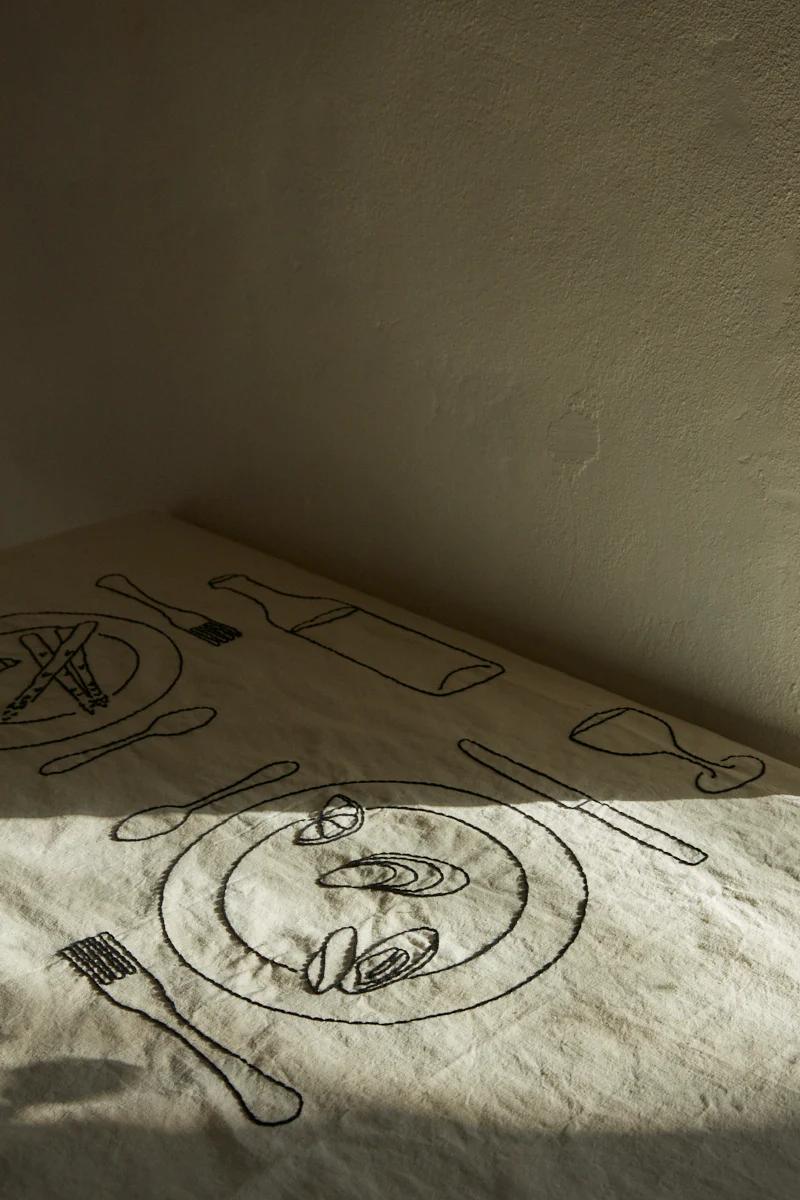 Tablecloth with black line drawings of place setting. Includes plates, utensils, wine glass, bottle, mussels.