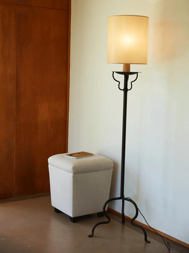 Minimalist corner with Balthus Lamp, beige shade. White ottoman with magazine nearby. Wooden wall, light floor.