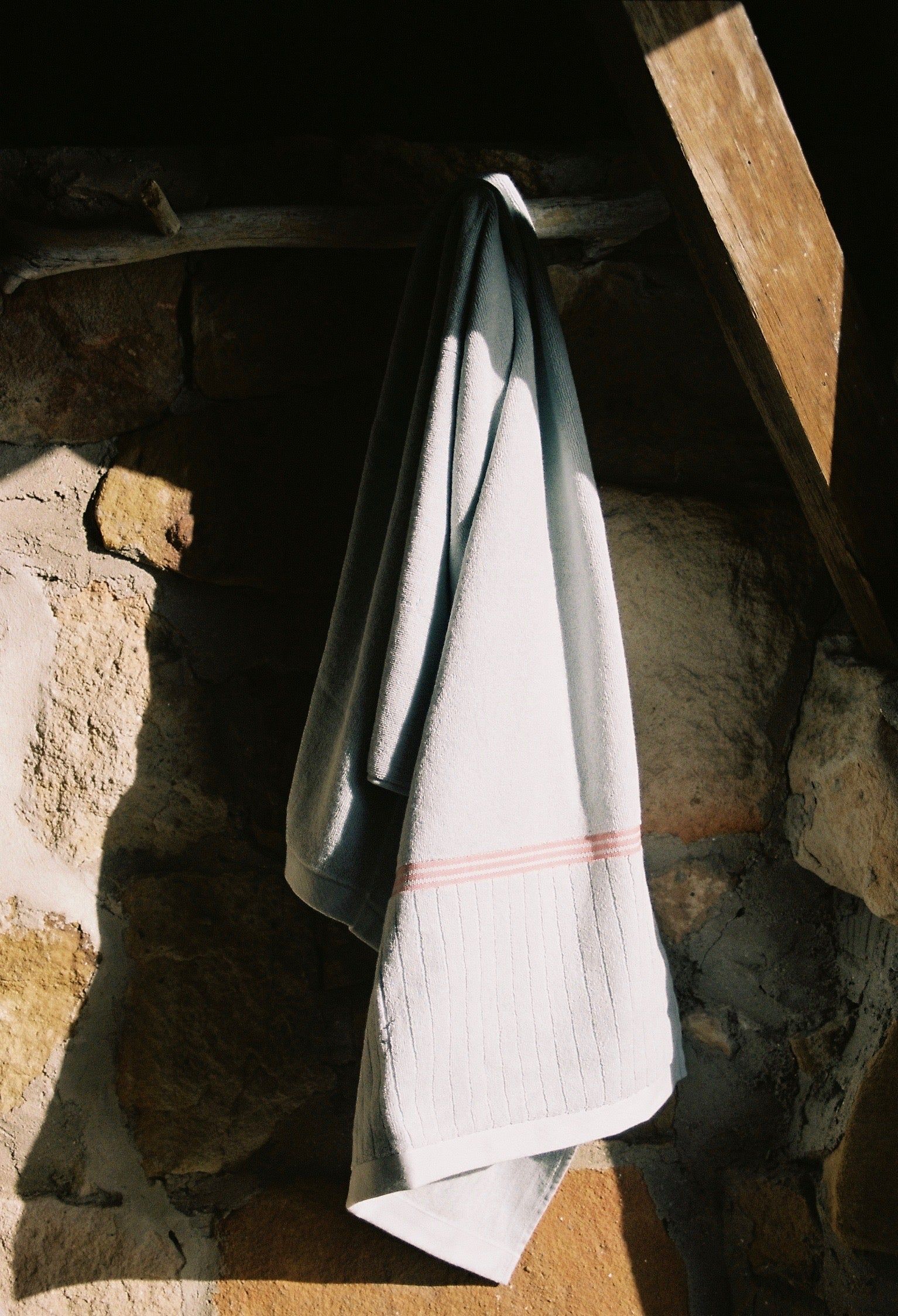 A white towel with a pink stripe hangs from a wooden branch on a stone wall, bathed in sunlight, creating a warm seaside cottage atmosphere.