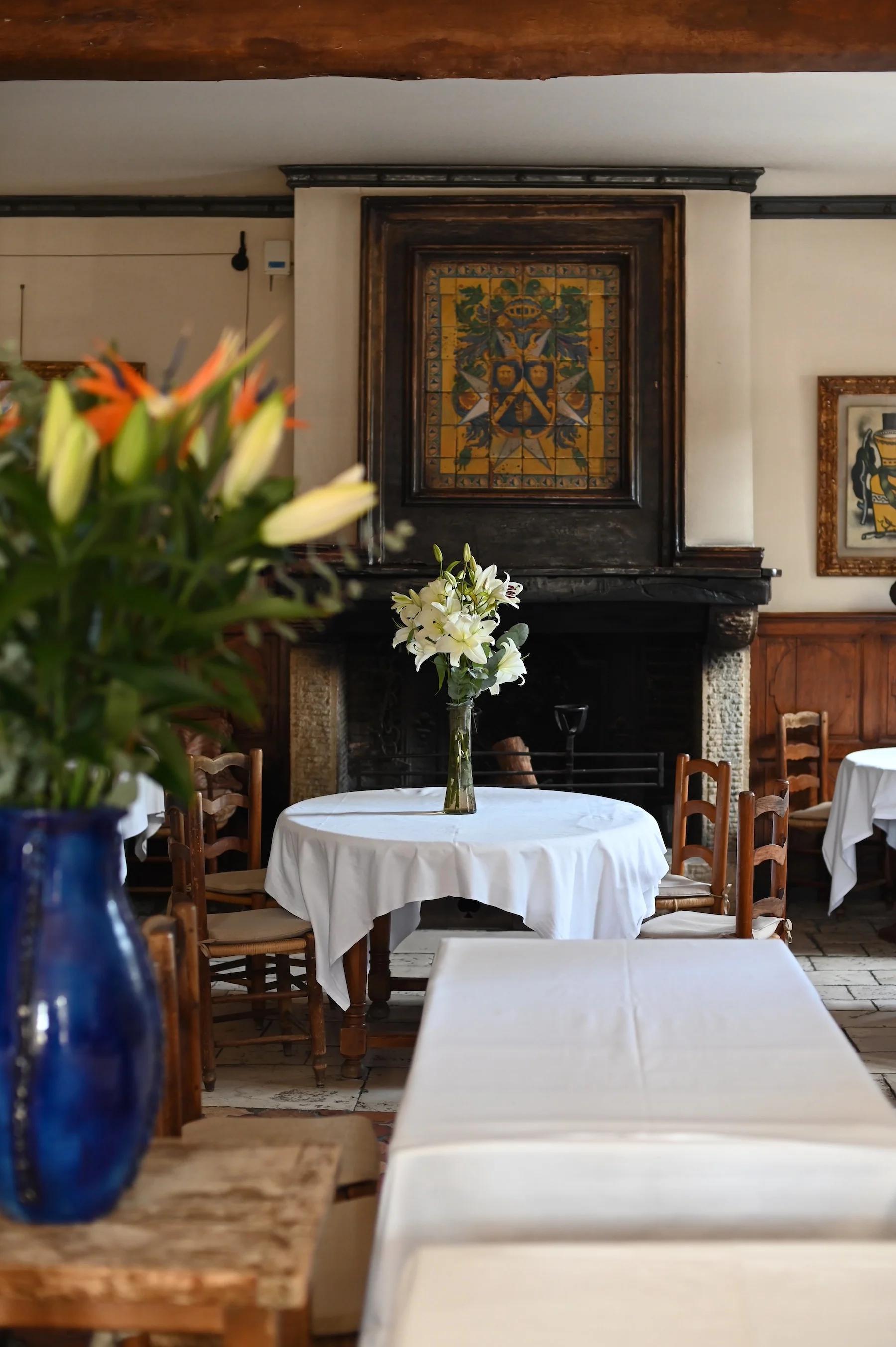 Cosy dining room with wooden tables and white tablecloths. White flowers in vases adorn tables. Ornate wall decoration above fireplace in background.