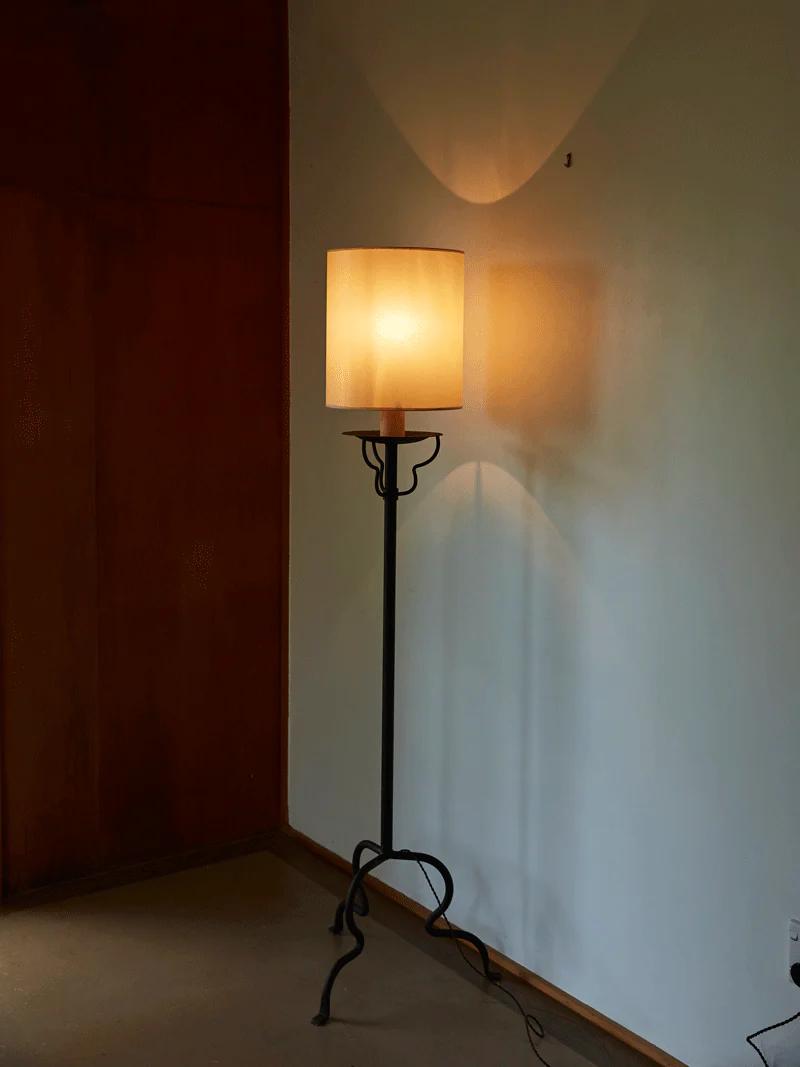 Balthus Lamp with beige shade against white wall. Black metal stand, three legs. Warm light casting shadows.