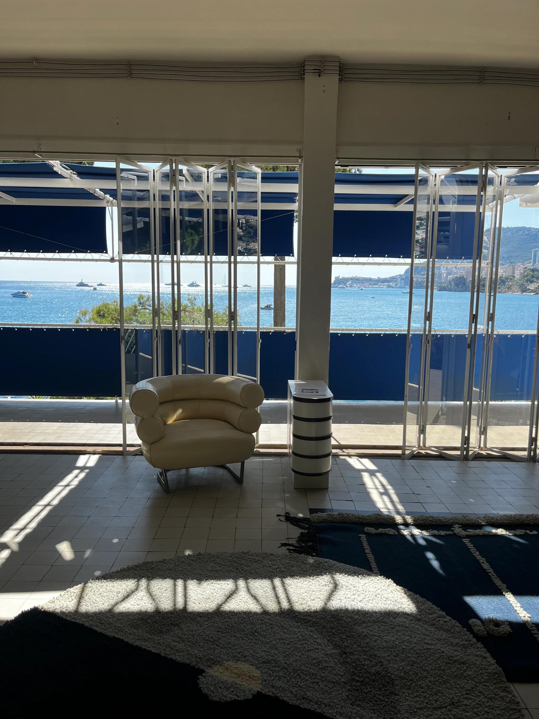 Modern lounge area with white armchair and side table under large windows. Ocean view visible. Round rug on floor.