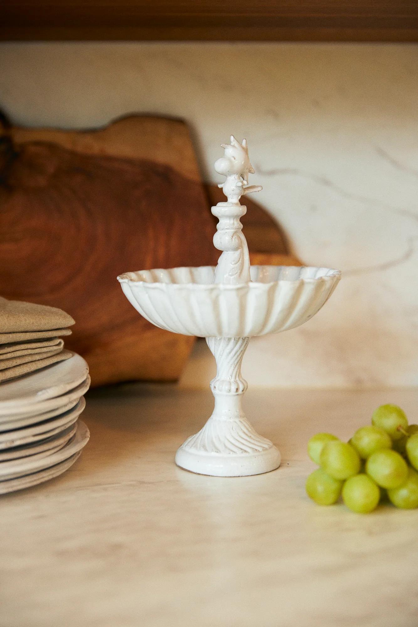 White ceramic pedestal dish with bird figure on marble counter. Stacked plates and wooden cutting board nearby. Green grapes visible.