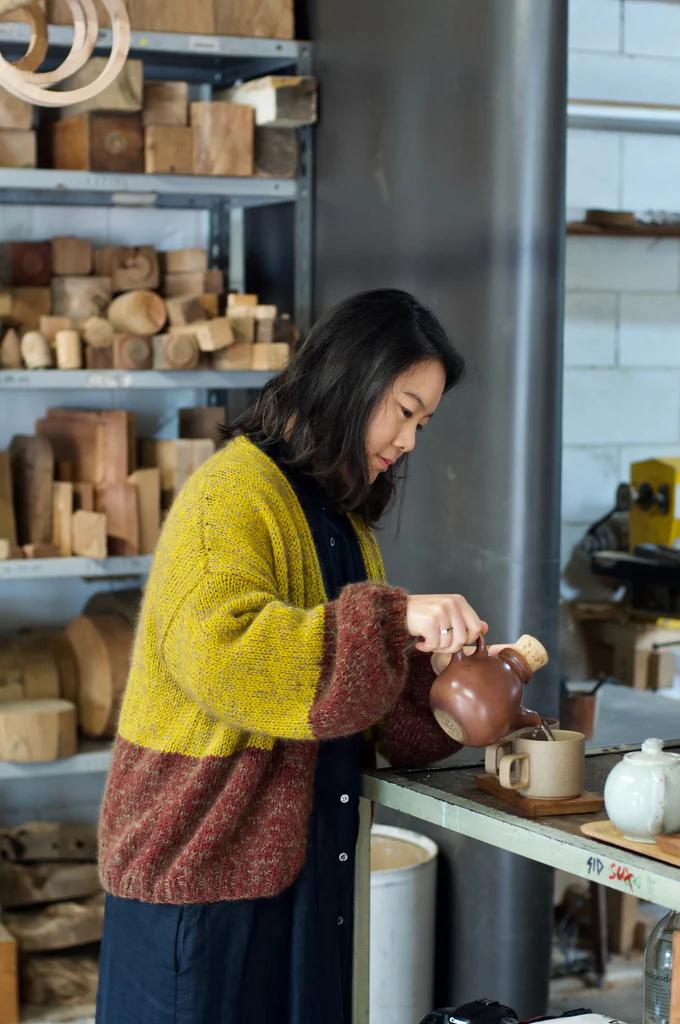 Dark-haired person in yellow-brown sweater pours from teapot at workbench. Background shelves display NZ wood and pottery pieces.