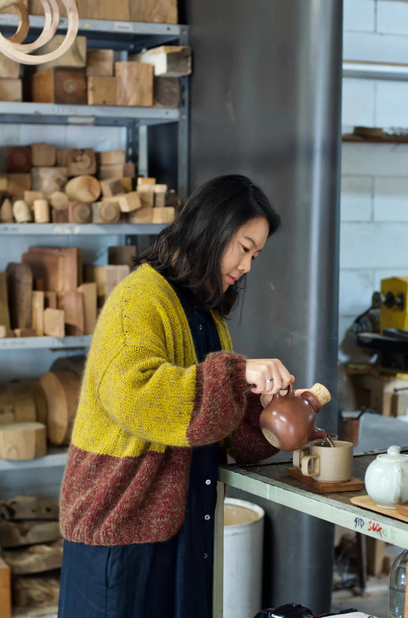 Dark-haired person in yellow-brown sweater pours from teapot at workbench. Background shelves display NZ wood and pottery pieces.