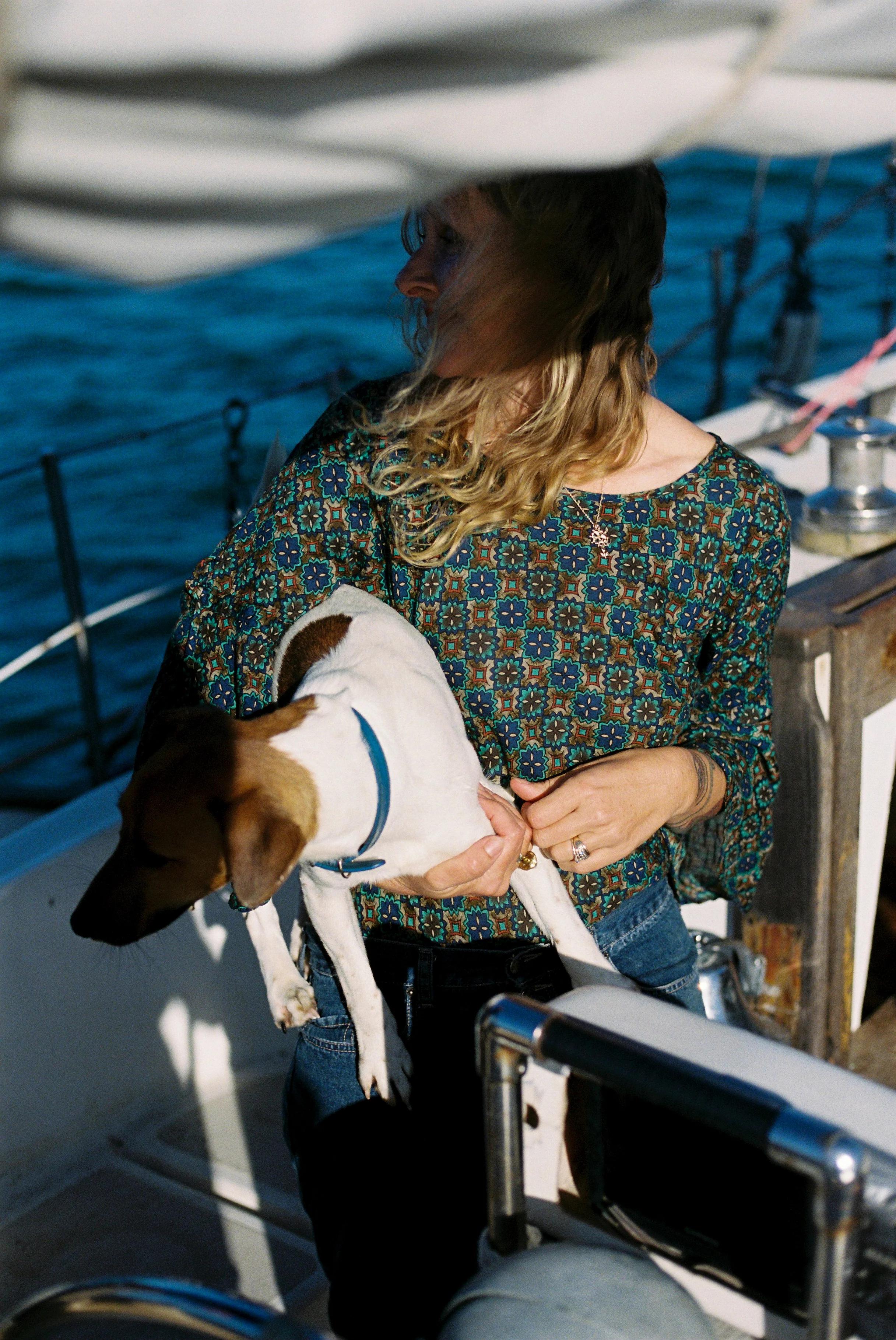 Person with blonde hair holding small dog on boat. Patterned shirt, water and railing visible.