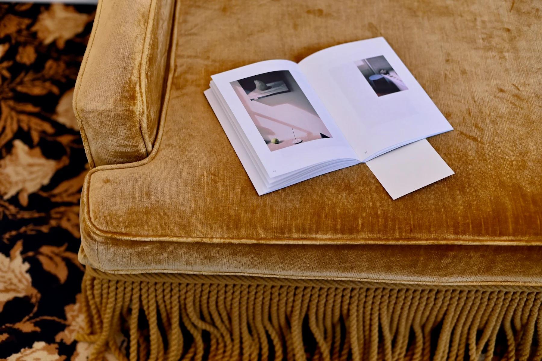 Open book on gold ottoman with tassels. Patterned black and white floral carpet beneath.