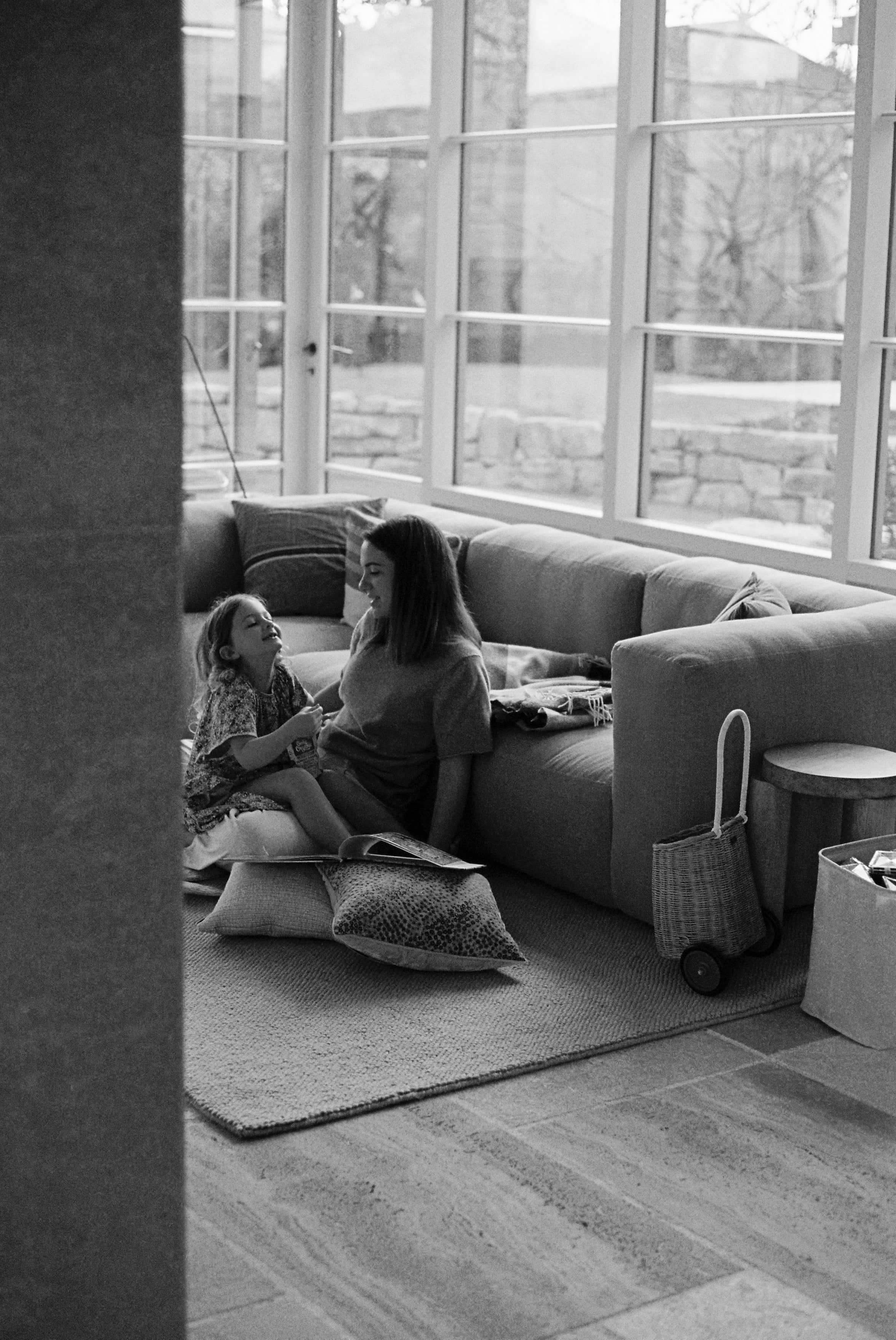 Black-and-white photo of woman reading to young girl on floor against couch. Cushions, blanket, and woven basket nearby.