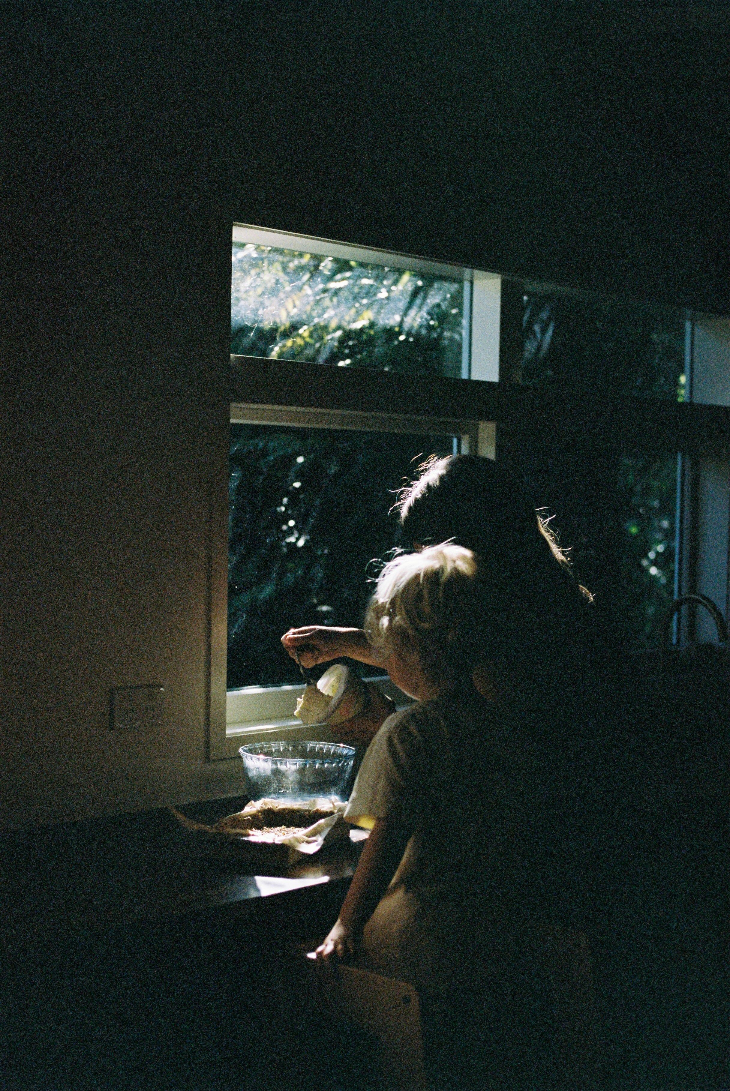 Adult and child by window. Adult pouring liquid, child watching. Sunlit, dimly lit scene.