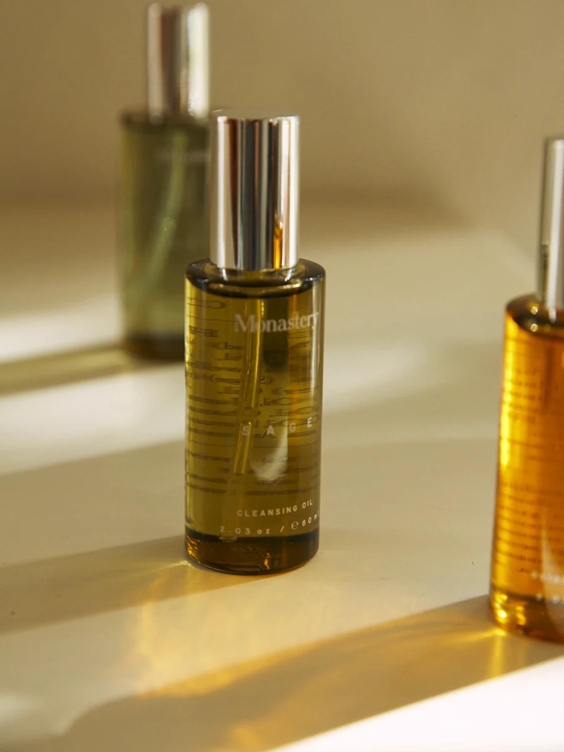 Monastery Sage Cleansing Oil bottle with silver cap on neutral background. Two blurry bottles behind, warm light casting shadows.