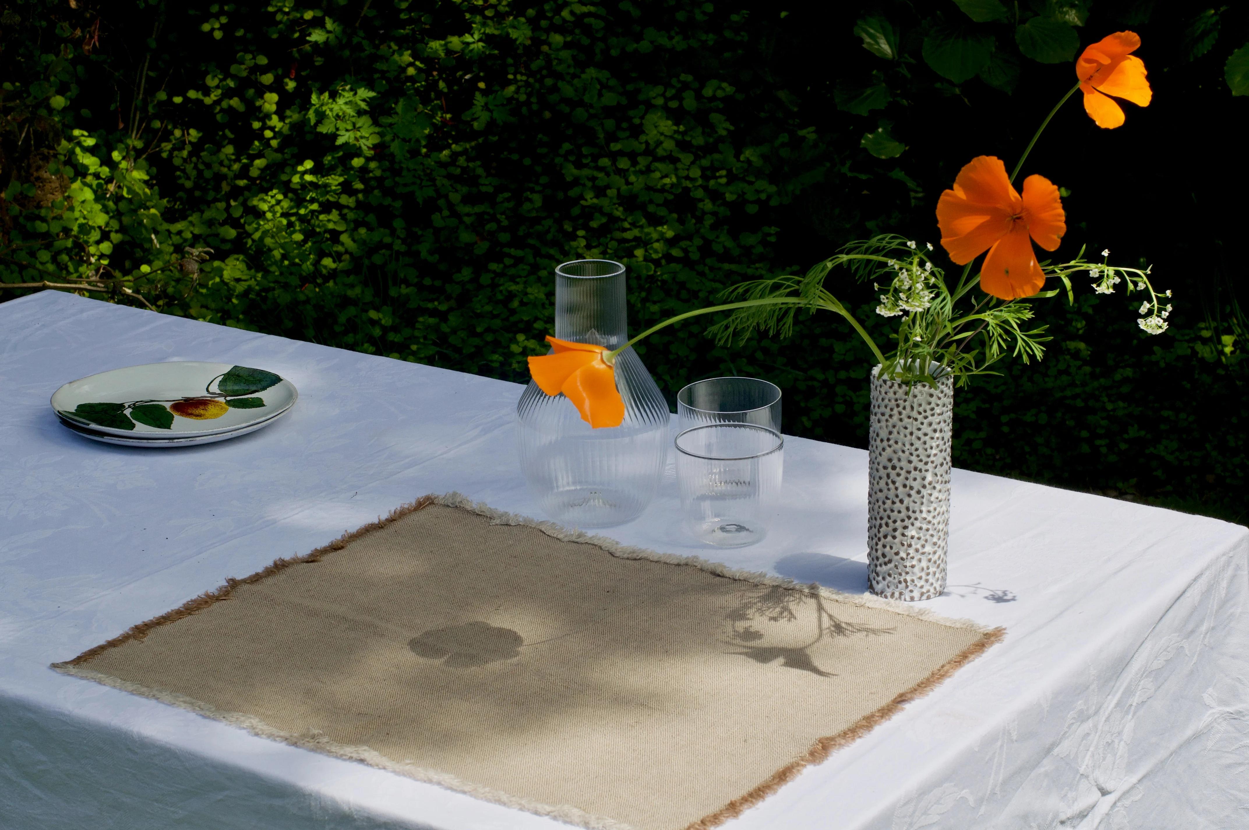 Outdoor table with elegant linens. Placemat, plate, glasses, carafe, and vase with flowers. Dappled sunlight creates serene ambiance.