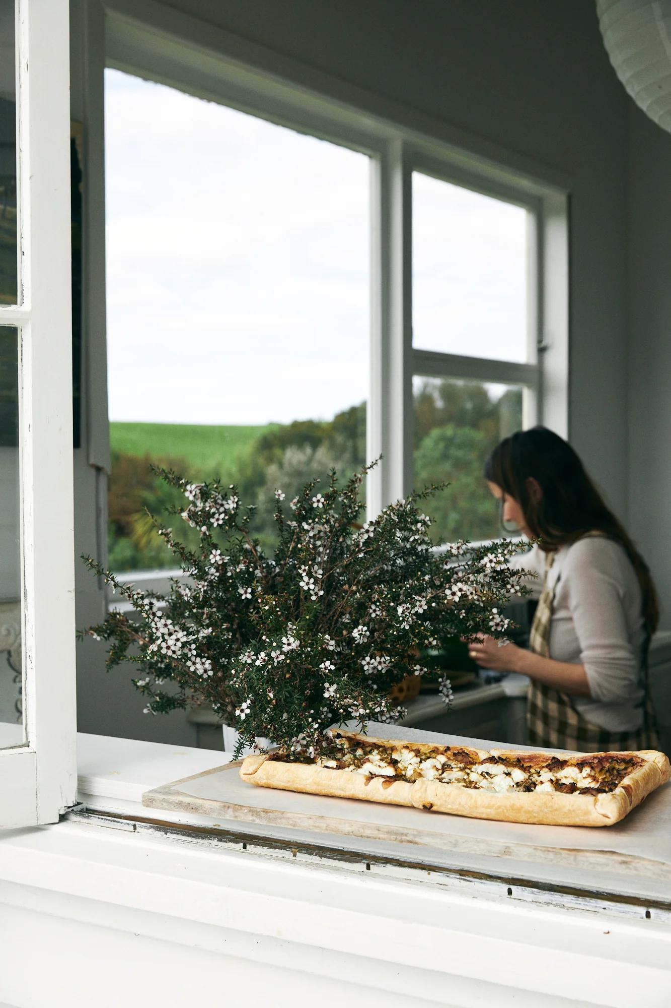 Savoury tart on wooden board by window. White flower vase. Woman in plaid apron cooking in background.