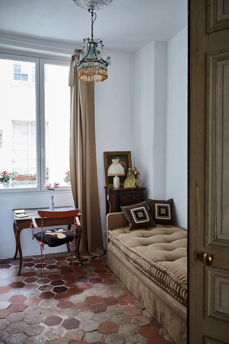 Elegant room with beige daybed and patterned pillows. Antique desk near curtained window, vintage chandelier, and decorative items evoke Parisian charm.