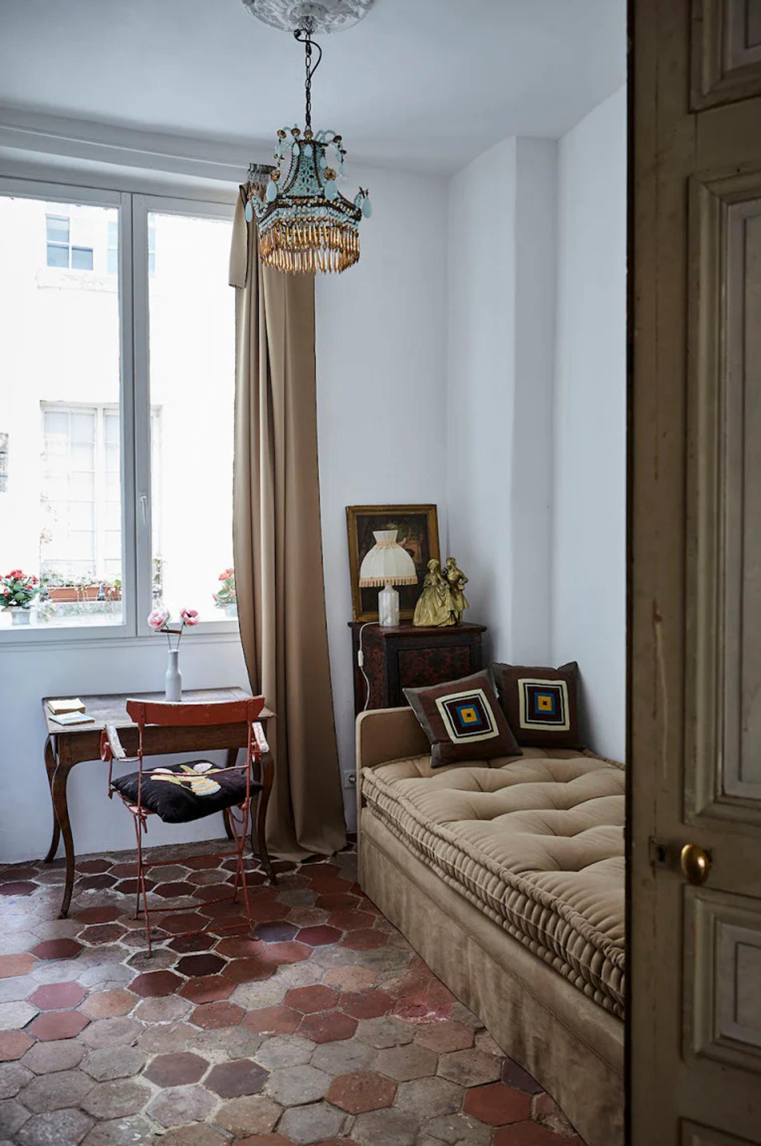 Elegant room with beige daybed and patterned pillows. Antique desk near curtained window, vintage chandelier, and decorative items evoke Parisian charm.