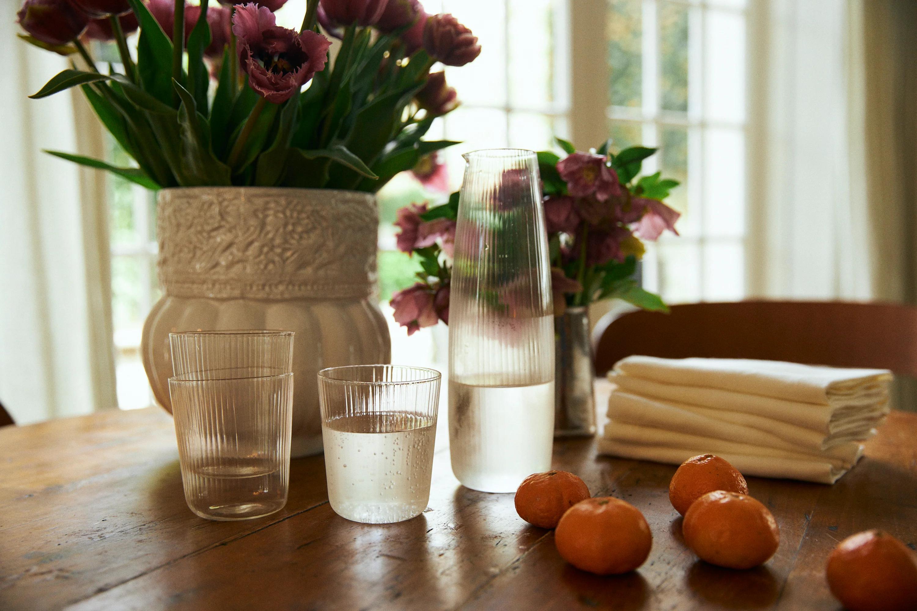 Table with water glasses, carafe, and mandarins. Vases with flowers and stacked napkins in background. Sunlight through curtains.