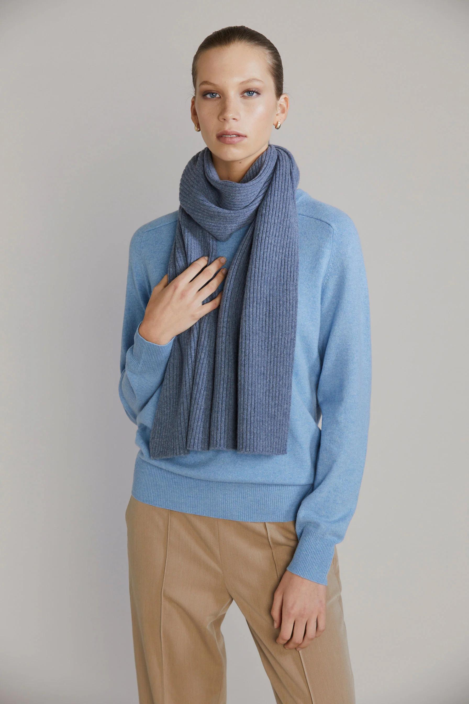 Person with tied-back hair wears light blue sweater, dark blue scarf, and beige pants. Hand touching scarf.