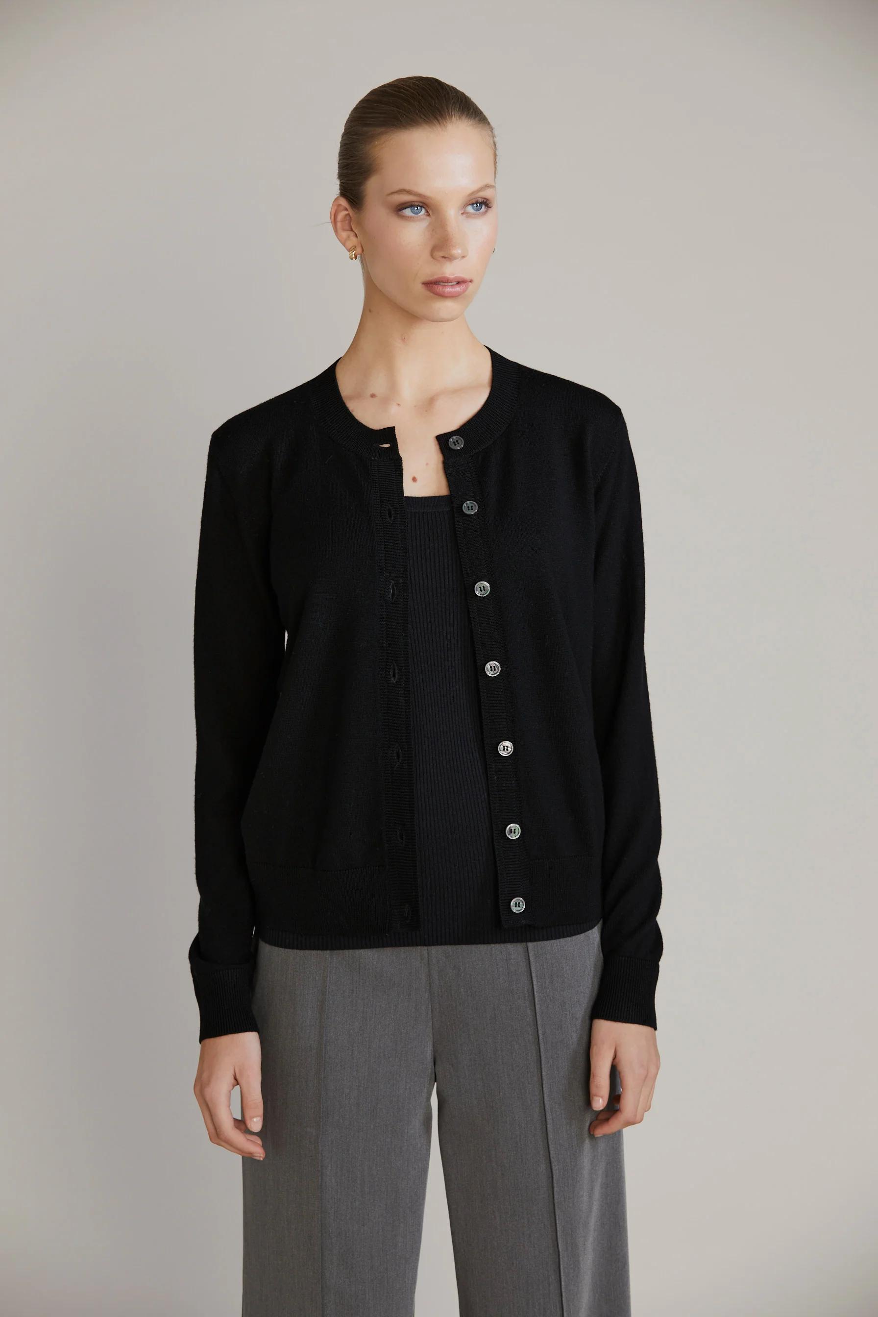 Person in black cardigan over black shirt and grey pants. Looking slightly to side. Plain background.