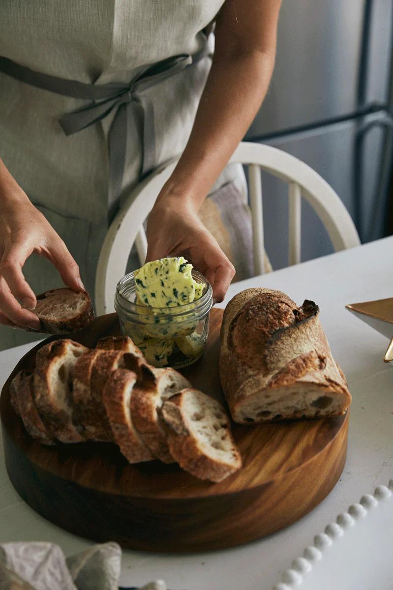 A person wearing a gray apron slices rustic bread on a round wooden board with herb butter. The scene evokes the warmth of holiday baking and Tessuti family recipes.