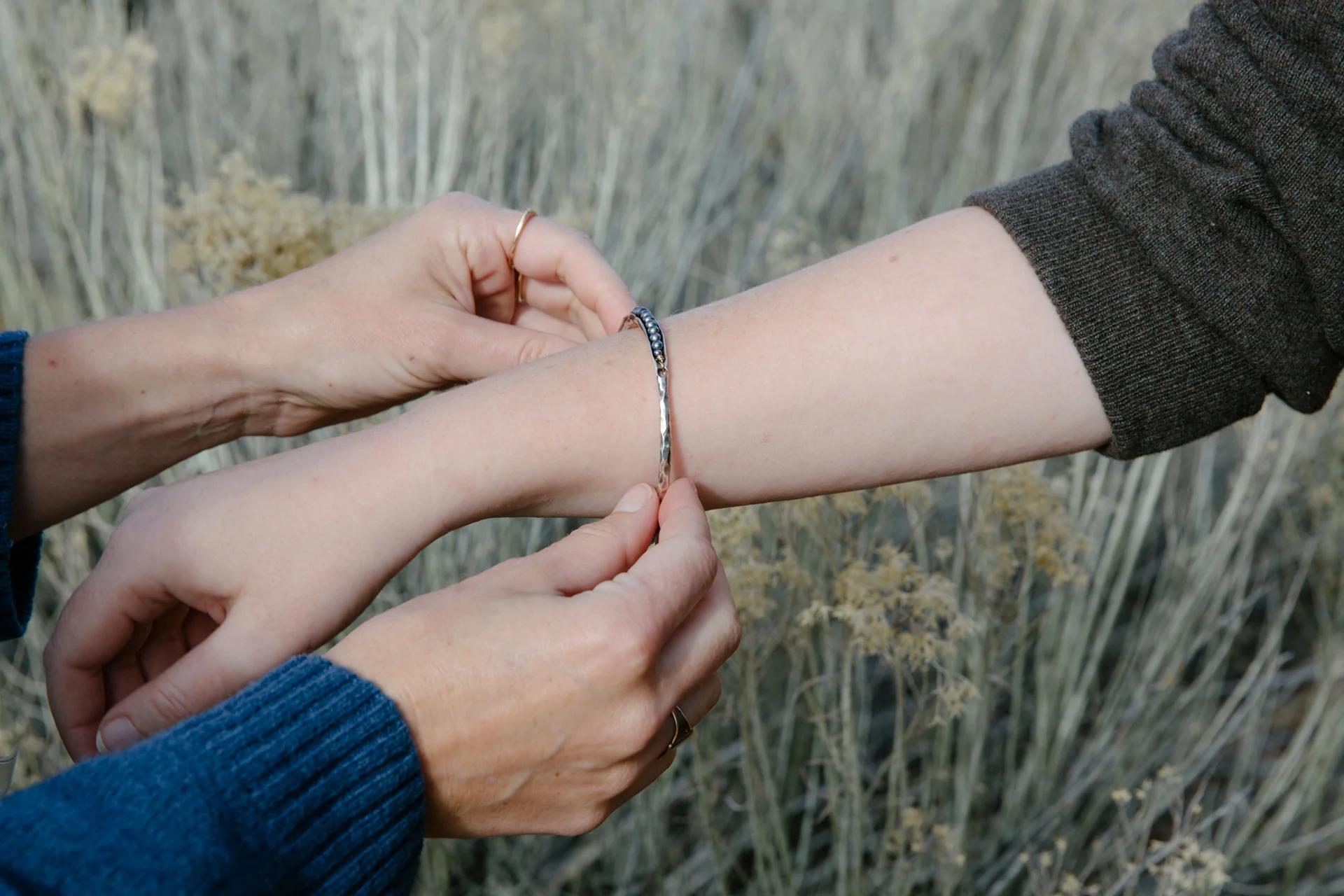 Hands fastening silver bracelet on wrist. Recipient in dark top, giver in blue sweater. Dried grass background.