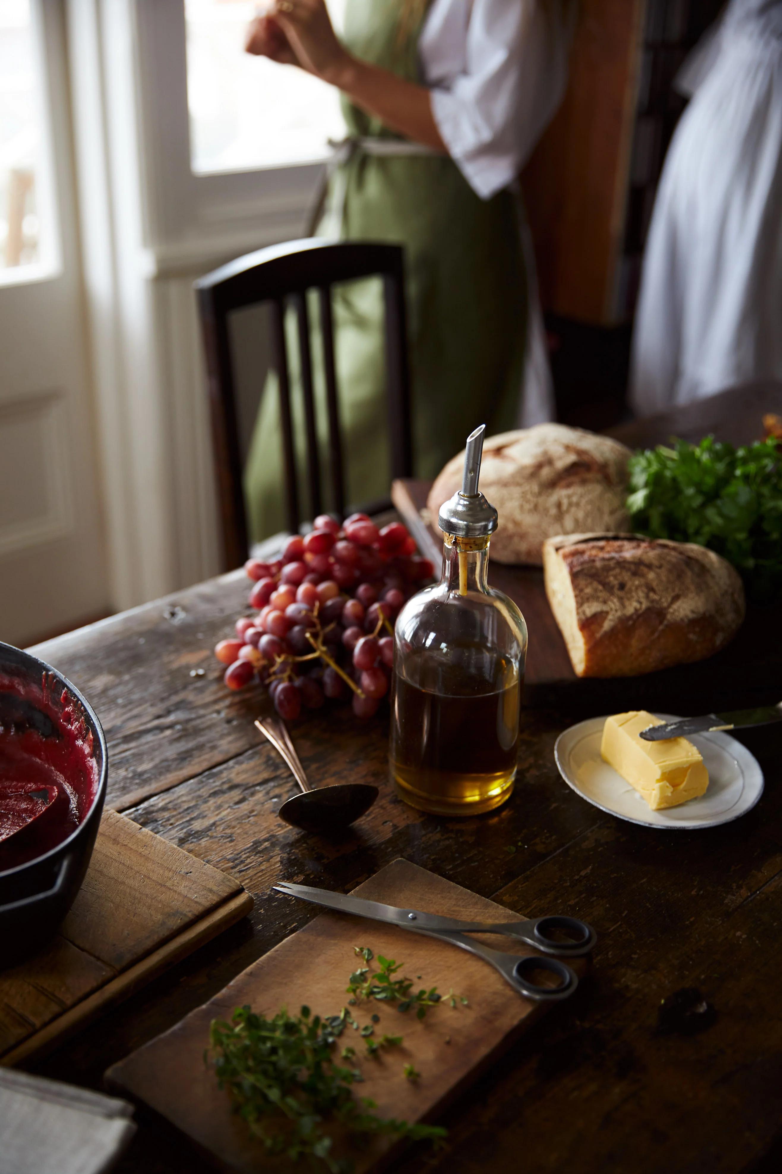 A rustic kitchen table with olive oil, bread, butter, grapes, and herbs. A person in a green apron stands by the window in the background.