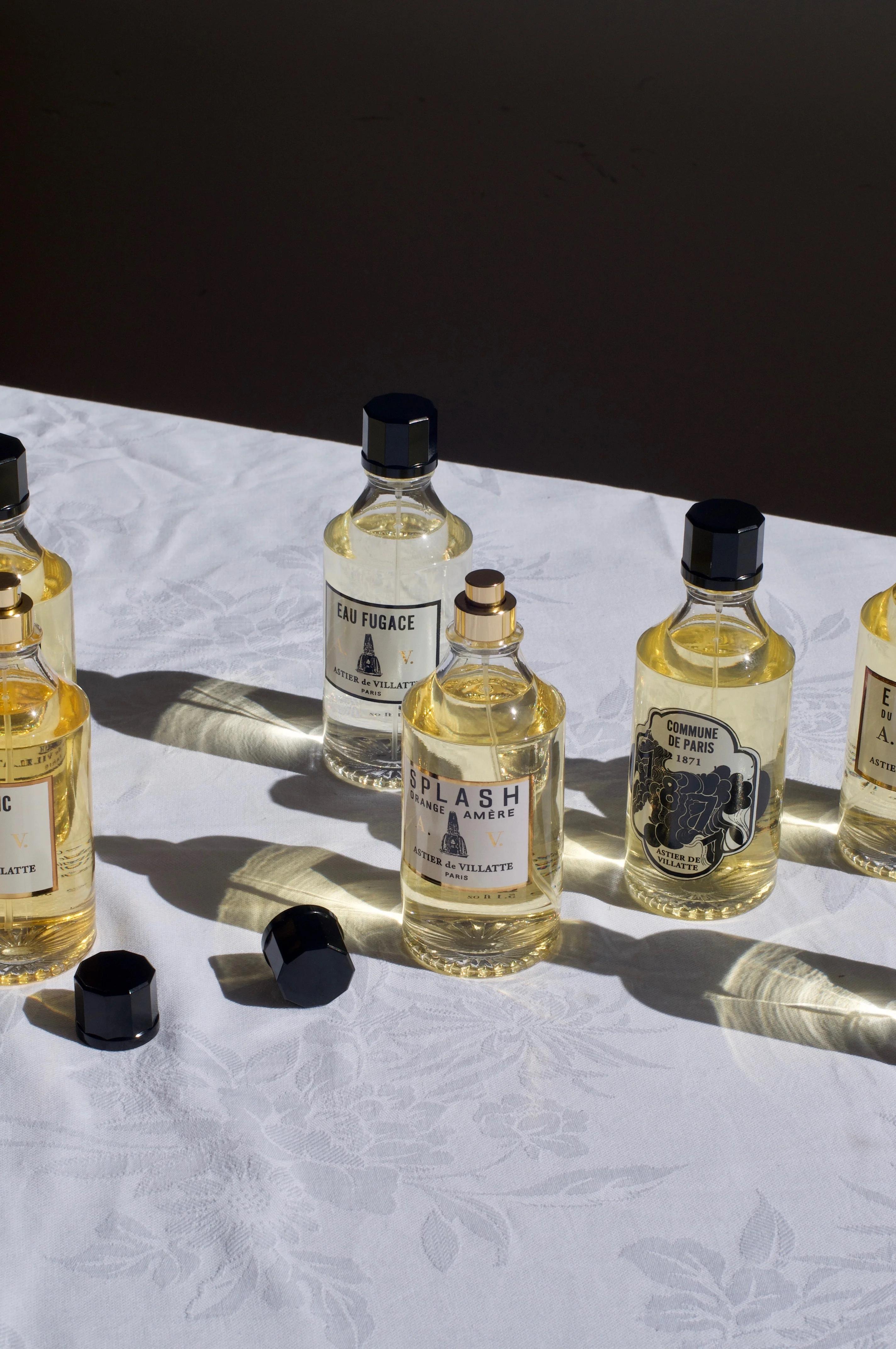 Multiple vintage-style perfume bottles with transparent yellow liquid on a white patterned surface. Sunlight casts shadows, and some caps are off, adding to the elegant scene.