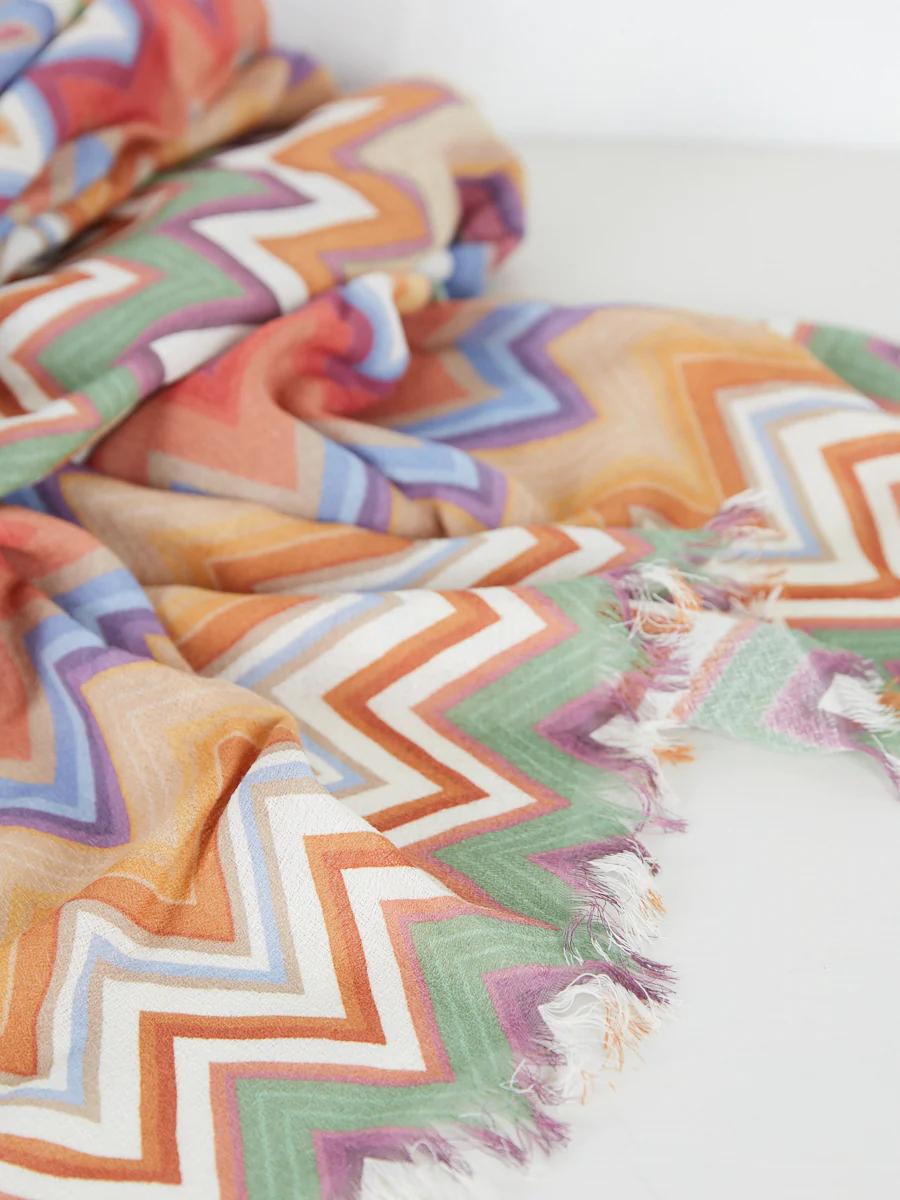 Close-up of colourful chevron fabric. Orange, green, pink, purple, white. Frayed edges. Light-coloured surface.