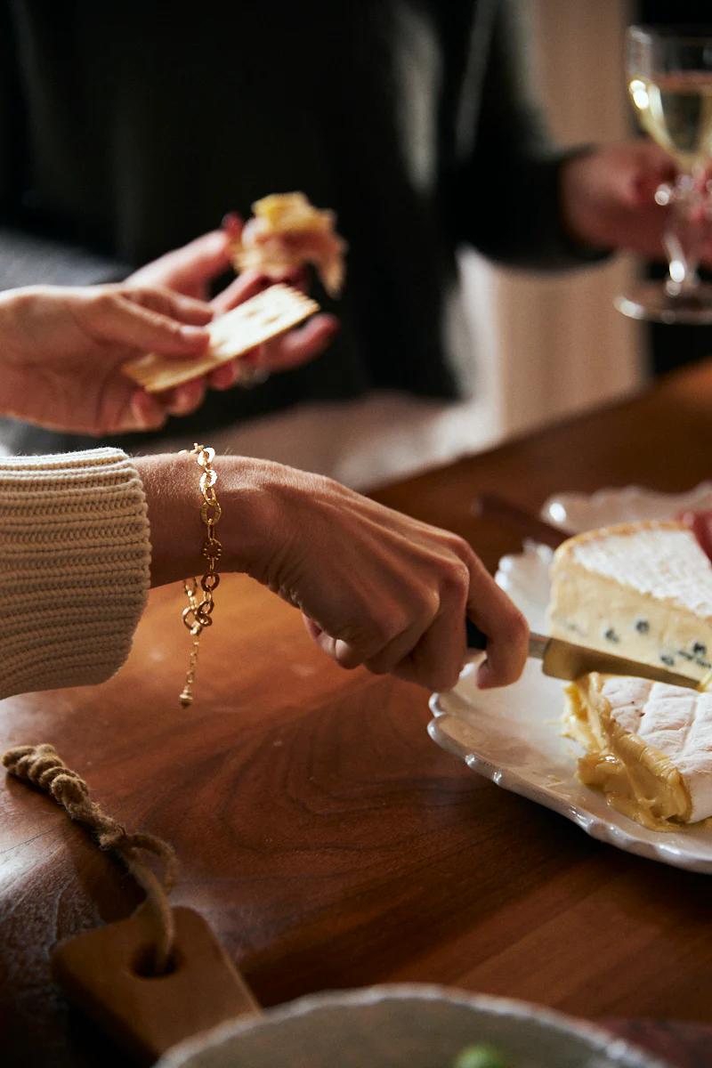 person is preparing food on a wooden table. Wearing a gold bracelet, they are cutting cheese on a white plate, and holding a square cracker in the other hand. Another person holds a glass of white wine in the blurred background. A cutting board with a rope handle is partially visible.