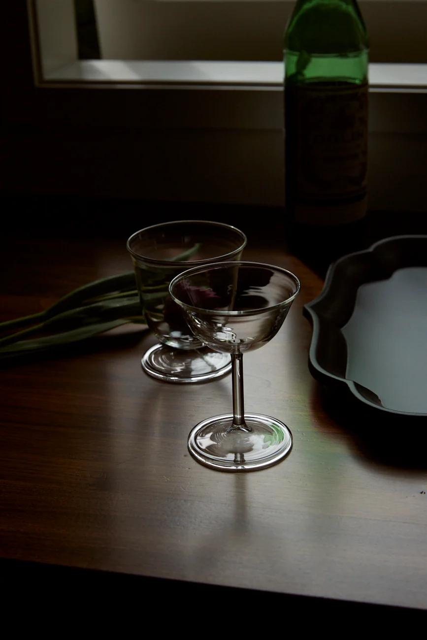 Two empty wine glasses sit near a black tray with a green bottle. Green stalks lie beside, showcasing Italian design in a dimly-lit setting.