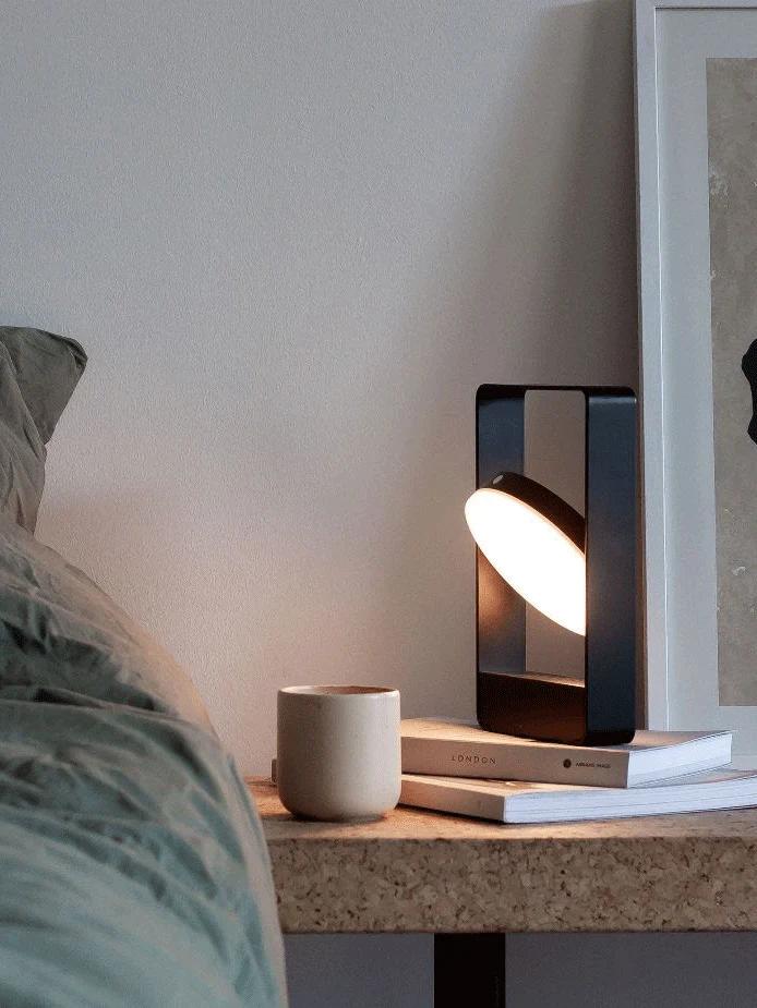 A cosy bedside table with a modern lamp, coffee mug, and stacked books, including one by Thich Nhat Hanh. Soft lighting creates a serene atmosphere.