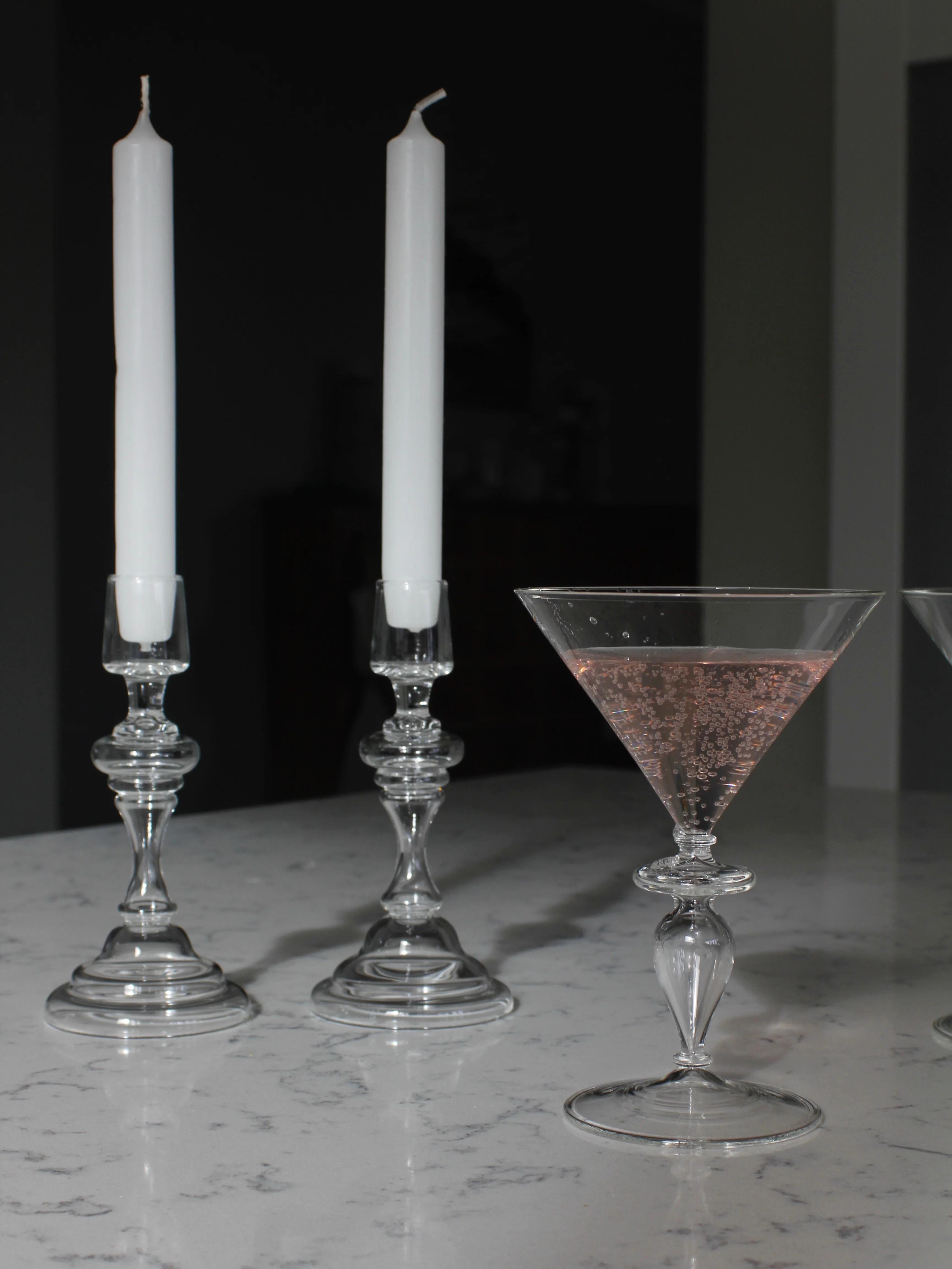 Two tall Astier de Villatte candles in glass holders on marble. Nearby, a stemmed glass holds pink fizzy drink. Scene exudes elegance and simplicity.