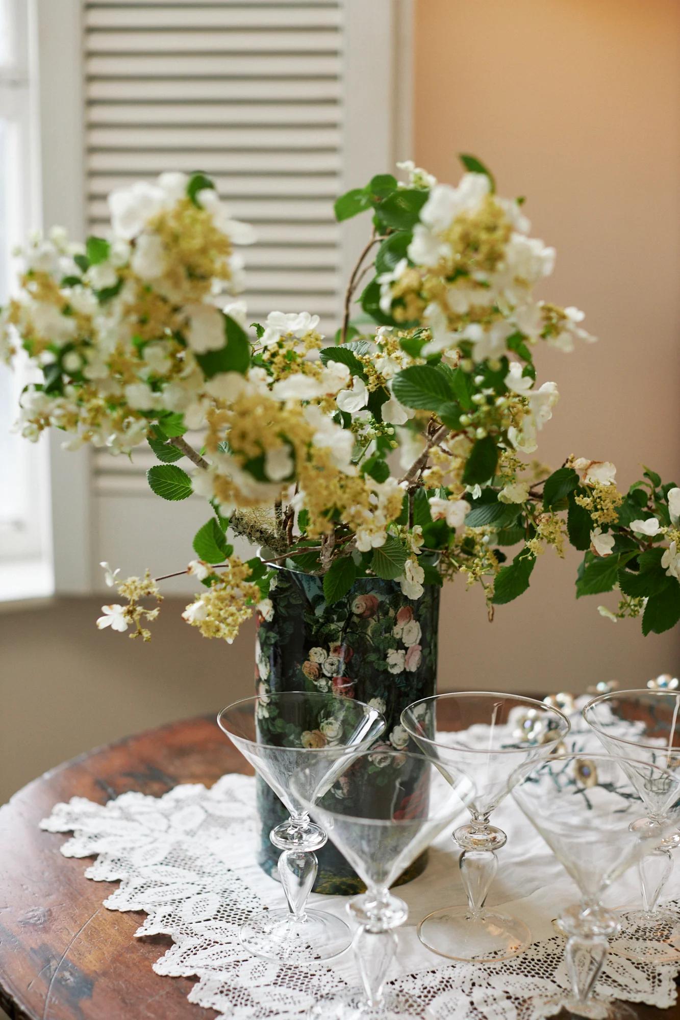 White flowers in a patterned vase on a lace-covered table. Empty cocktail glasses surround the centrepiece, with a louvered window in the background.