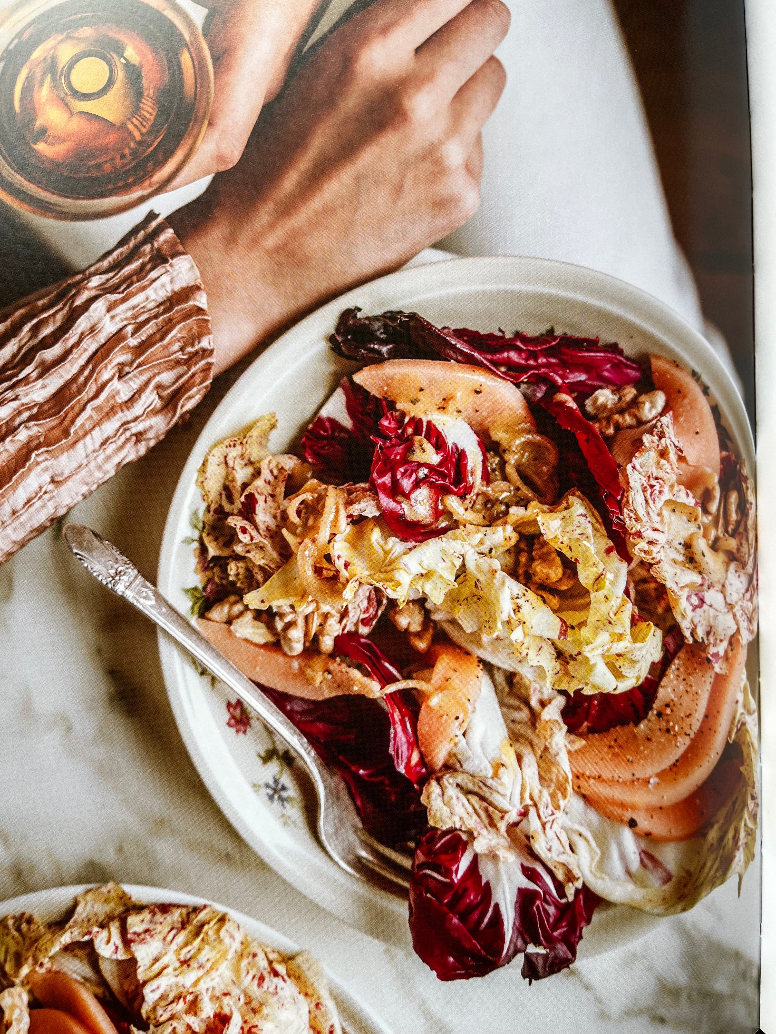 Person holds glass near colourful salad bowl. Salad includes radicchio, cabbage, and nuts. Patterned sleeve visible. Fork on bowl edge.