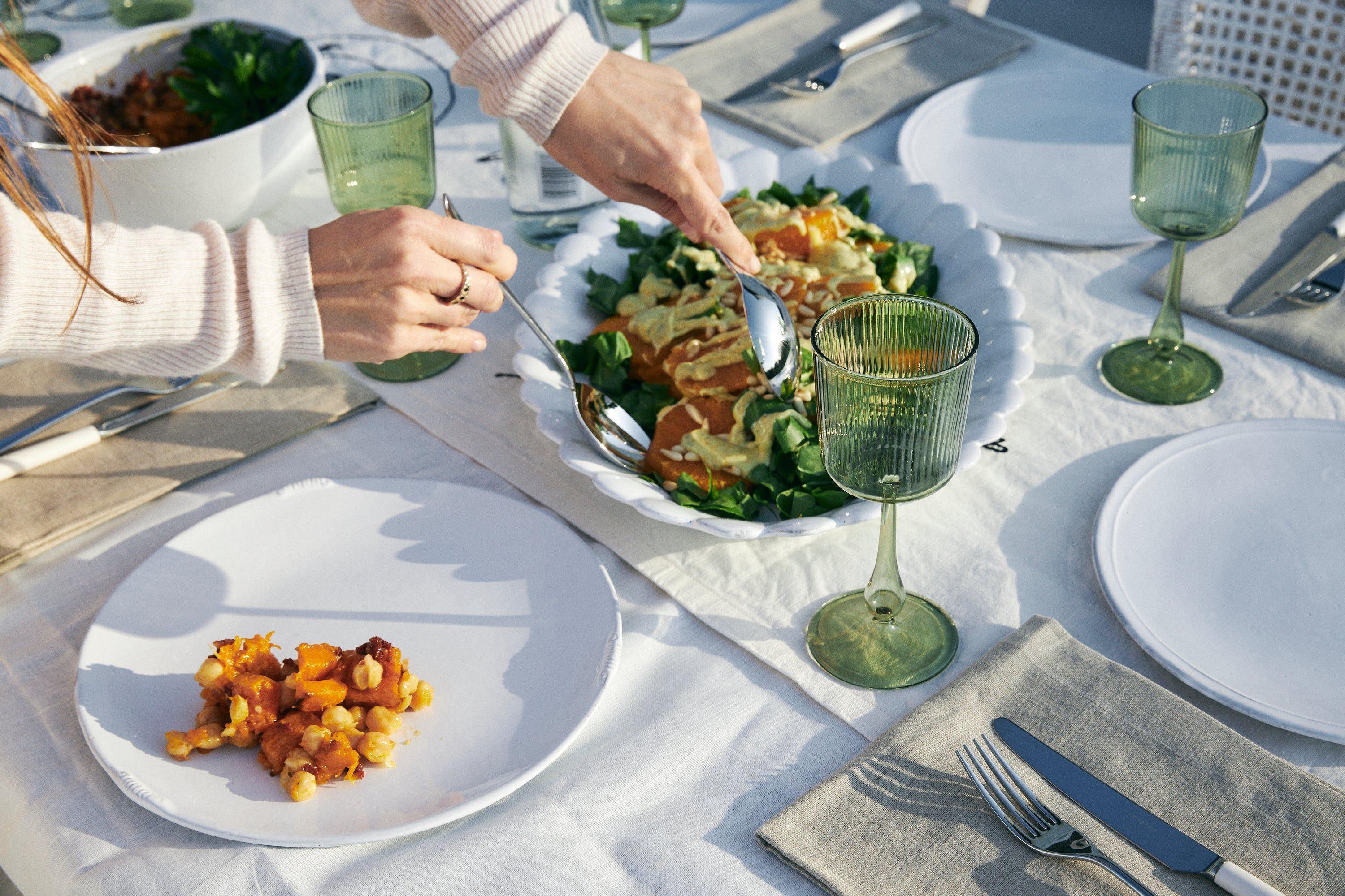 Person serving salad with tongs. Table with white cloth, green glasses, gray napkins, white plates.