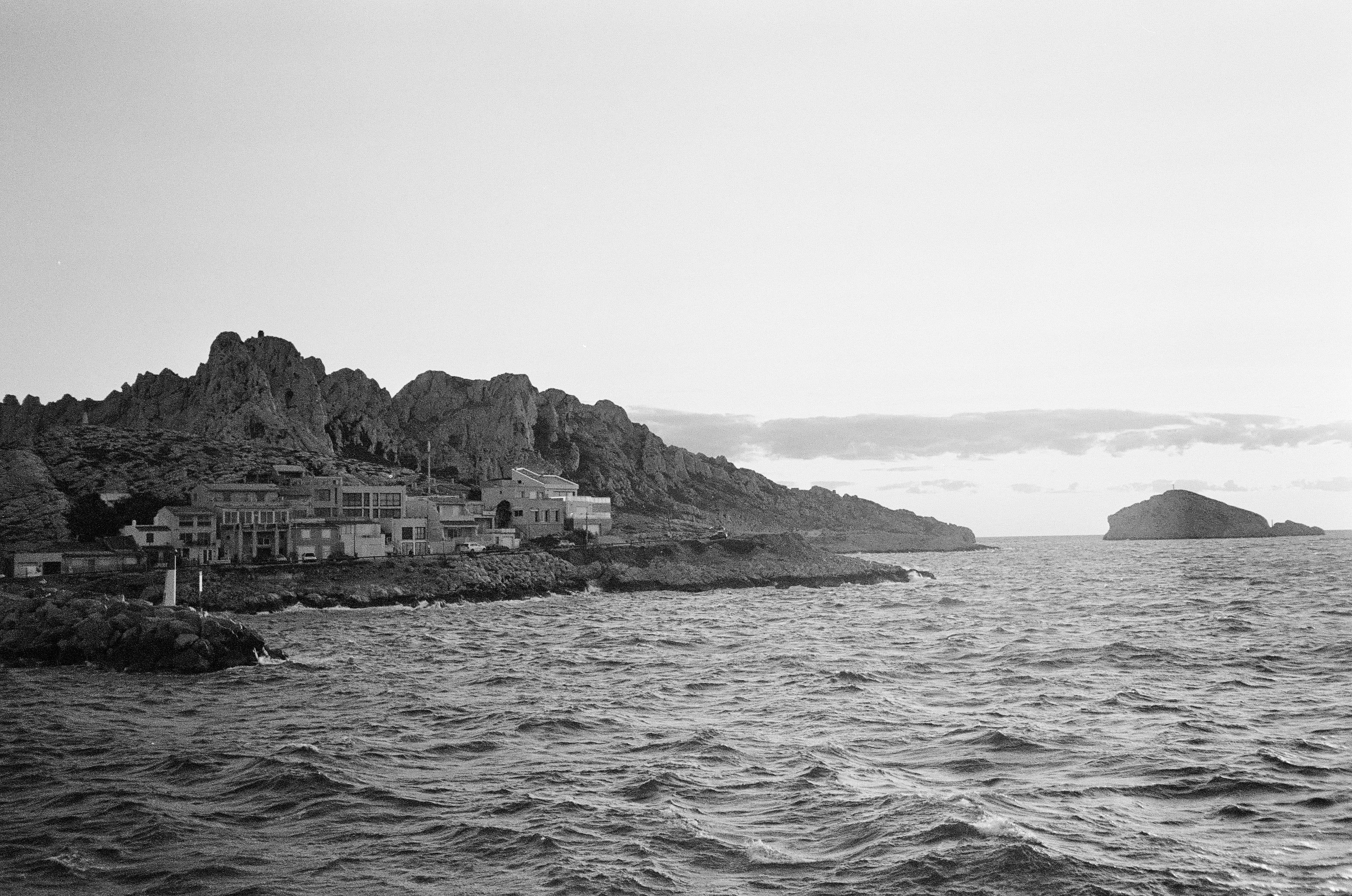 Grayscale coastal landscape with rocky cliffs, calm sea, and distant island. The scene evokes the tranquility of Marseille's shores.