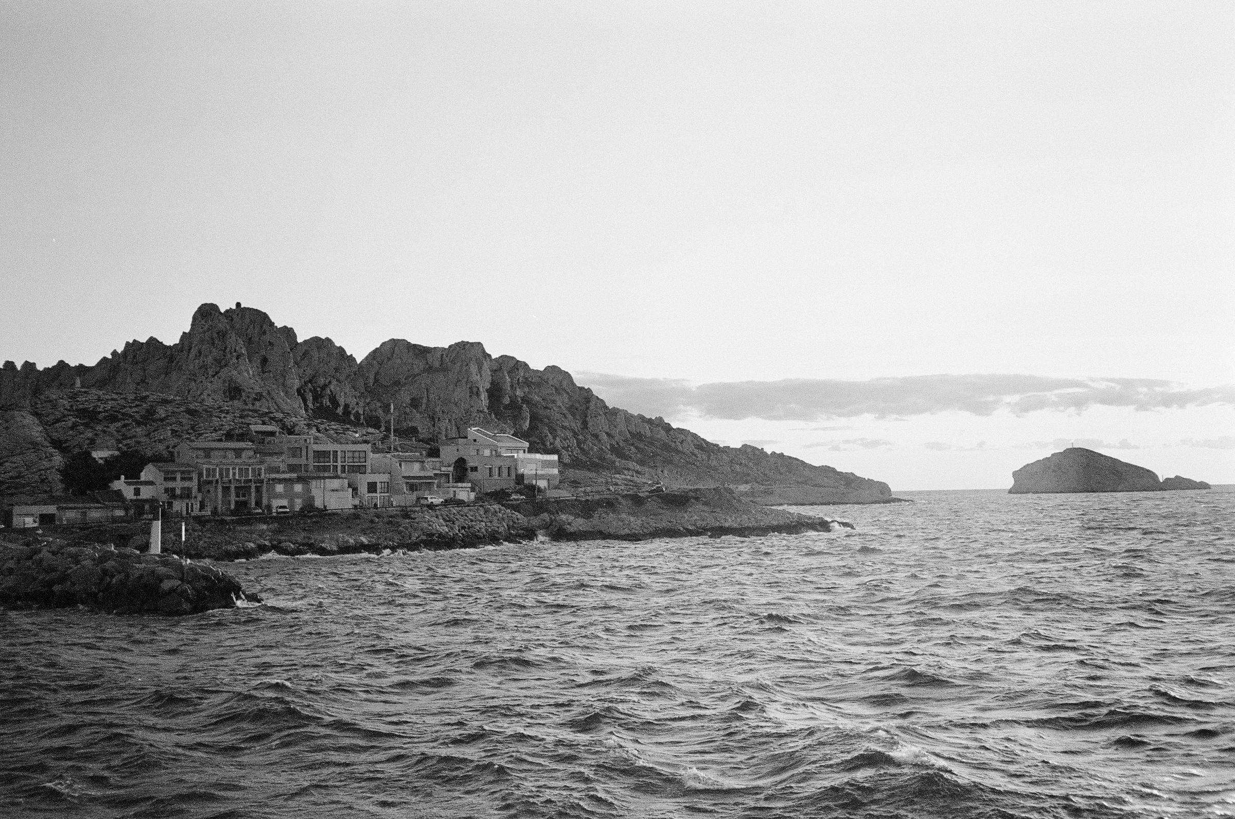 Grayscale coastal landscape with rocky cliffs, calm sea, and distant island. The scene evokes the tranquility of Marseille's shores.