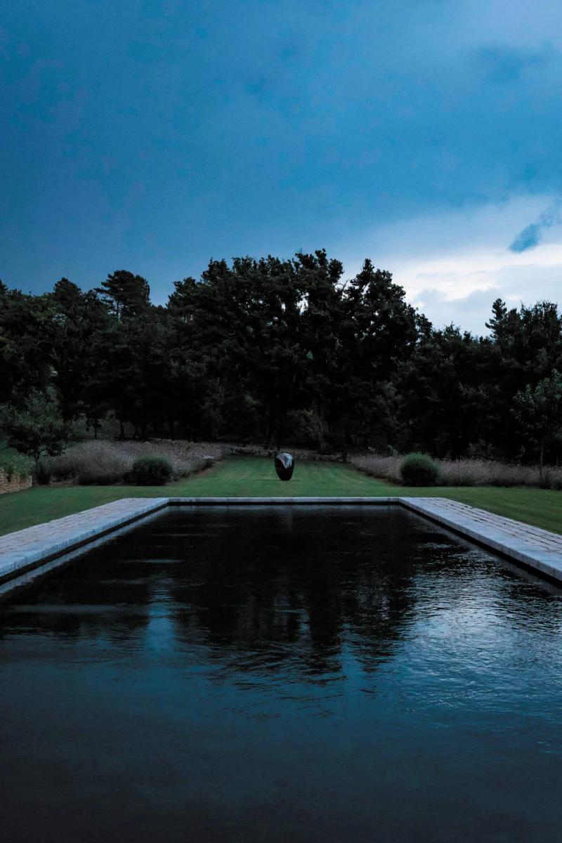 A reflecting pool bordered by stone paths at dusk. A dark sculpture stands on grass with trees behind, creating a serene Provençal atmosphere.