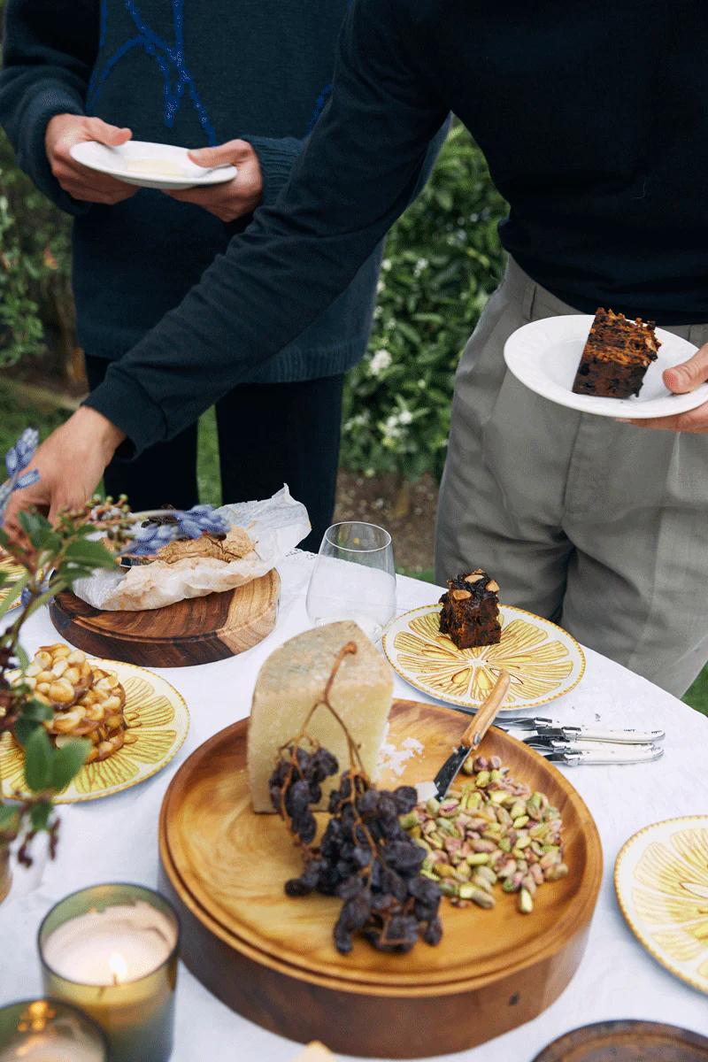 Two people serving themselves at outdoor table. Cheese, dried grapes, nuts, and Christmas cake visible.