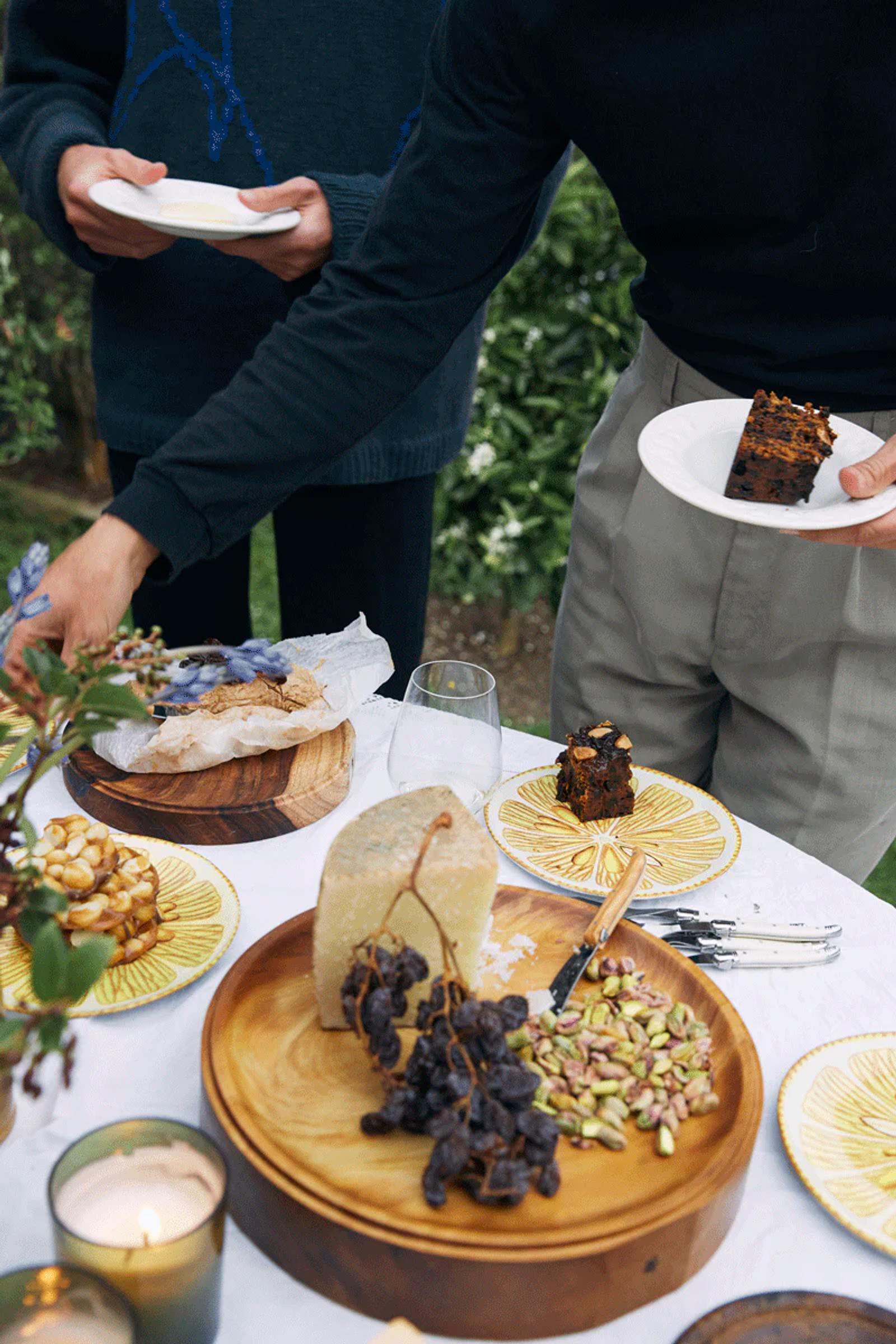 Two people serving themselves at outdoor table. Cheese, dried grapes, nuts, and Christmas cake visible.