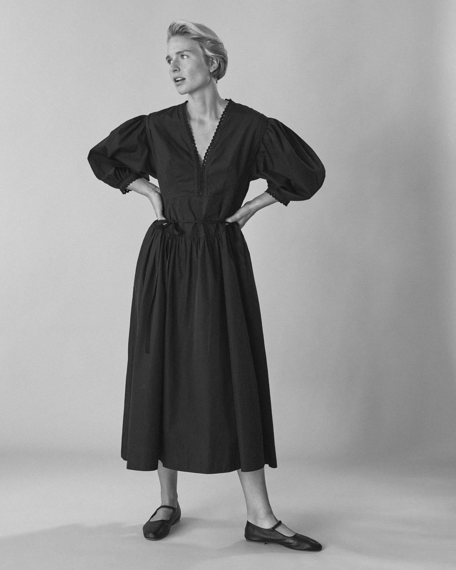 Person in long dark dress with puffy sleeves and V-neck. Hands on hips, short light hair. Light-coloured background.