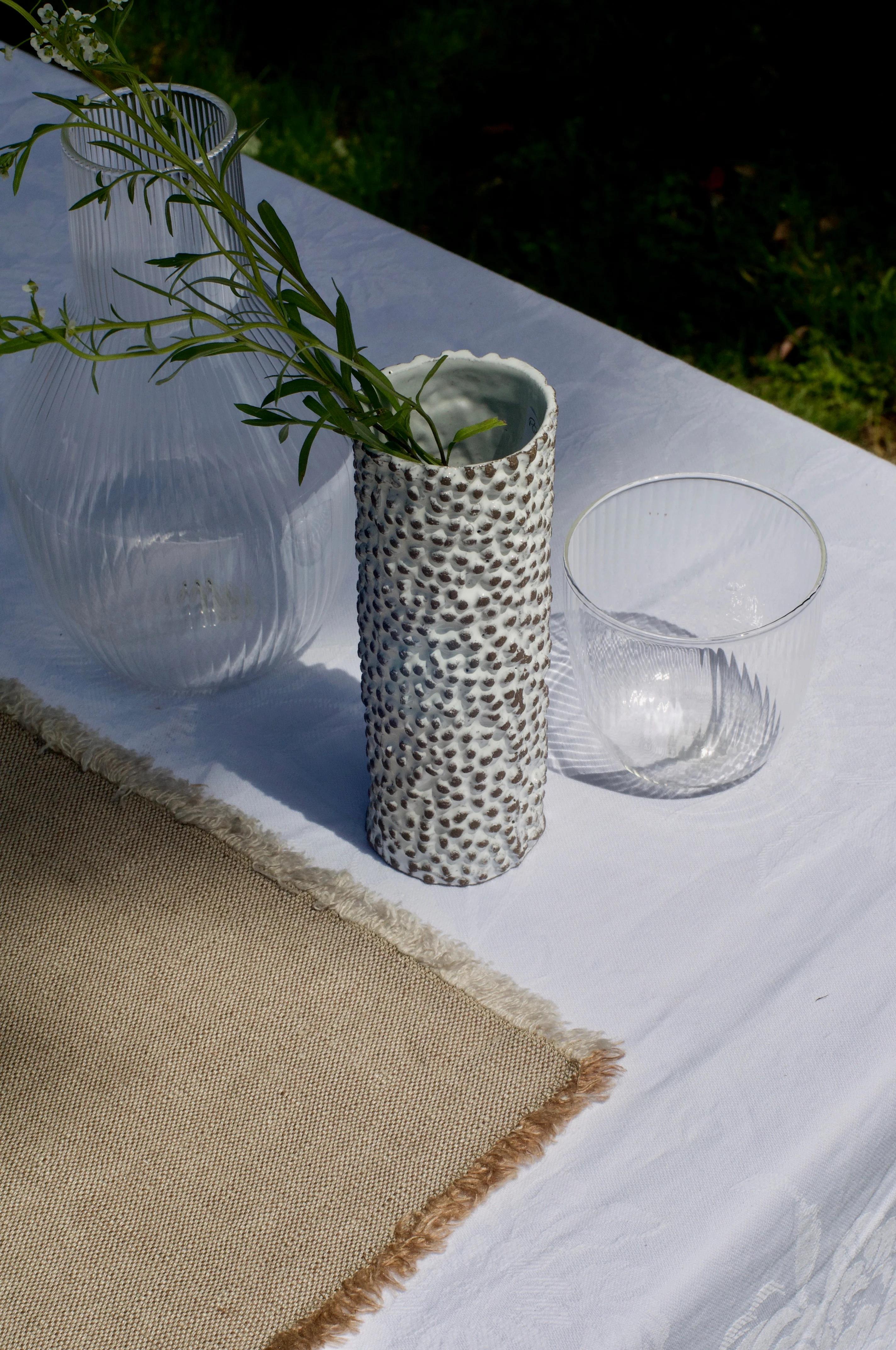 Outdoor table with white cloth. Ceramic vase, glass containers, and textured placemat visible. Italian brand table linens.