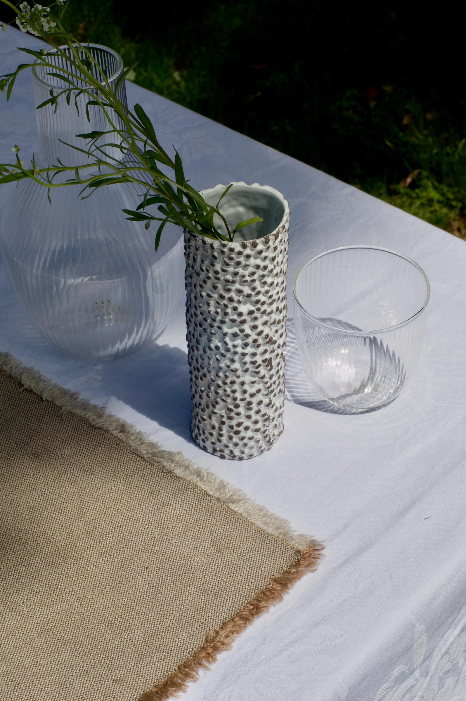 Outdoor table with white cloth. Ceramic vase, glass containers, and textured placemat visible. Italian brand table linens.