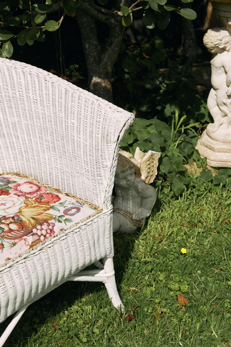 Partial view of white wicker chair with floral cushion on grass. Cherub statue and greenery create holiday garden scene.