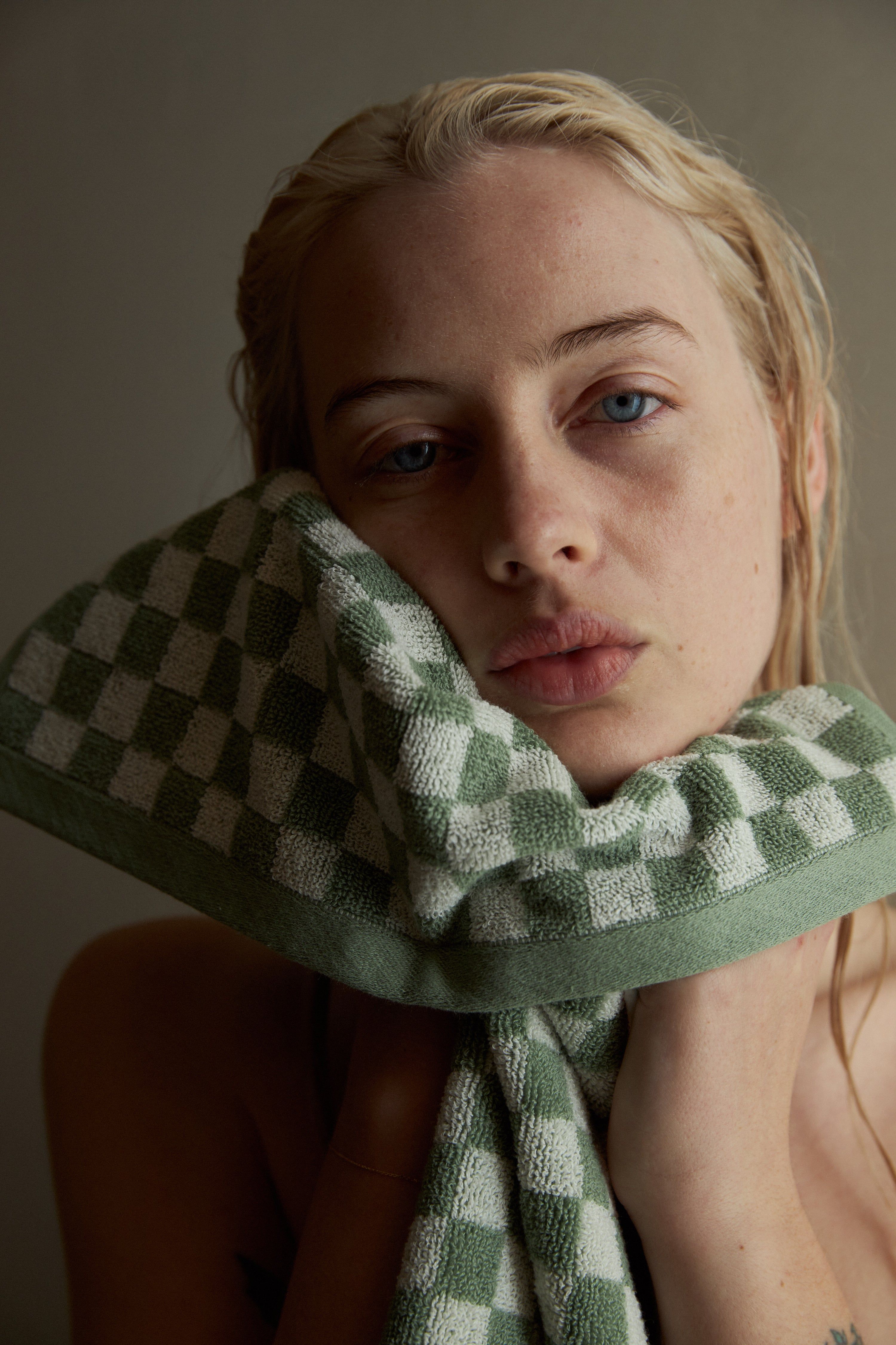 A person with wet hair holds a green and white checkered organic cotton towel to their face. The plain background focuses attention on the towel’s texture and craftsmanship.