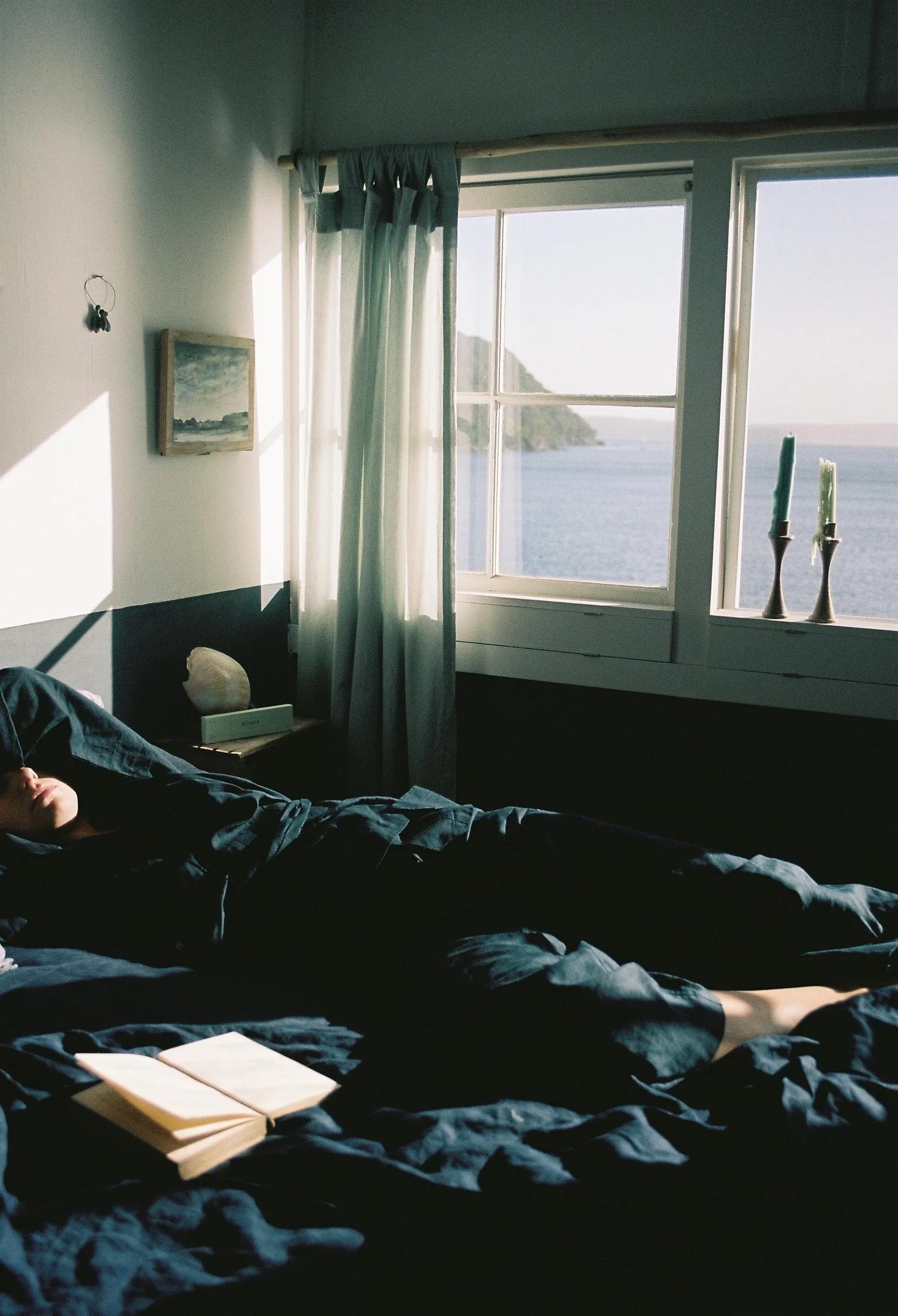 Sunlit bedroom with a person resting and an open self-care book. The window shows a calm sea and hills. Simple decor includes a painting and candles.