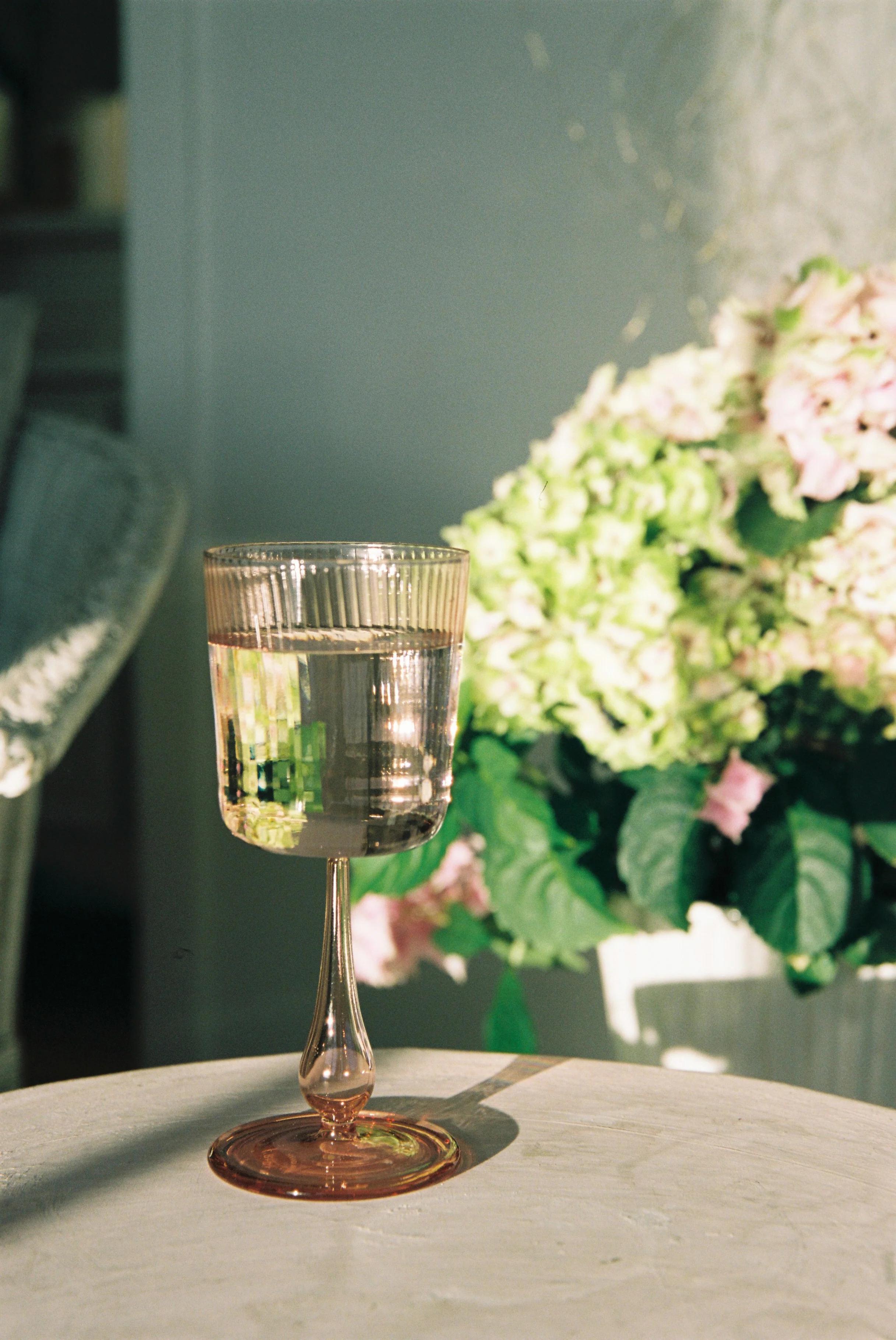 A pink-tinted vintage wine glass filled with white wine sits on a table. In the blurred background, green and pink flowers are bathed in sunlight, perfect for Mother’s Day pampering.