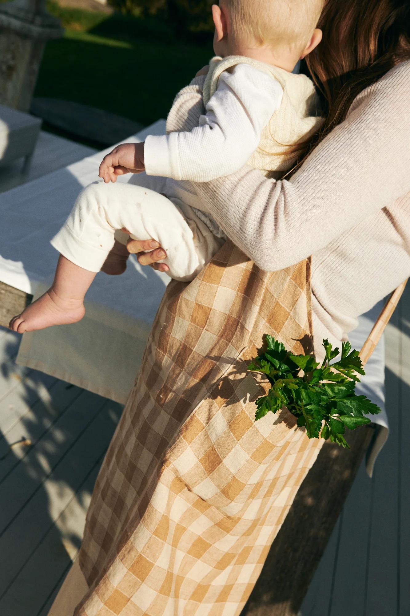 Person carrying baby, wearing checked apron with parsley. On wooden deck, greenery in background.